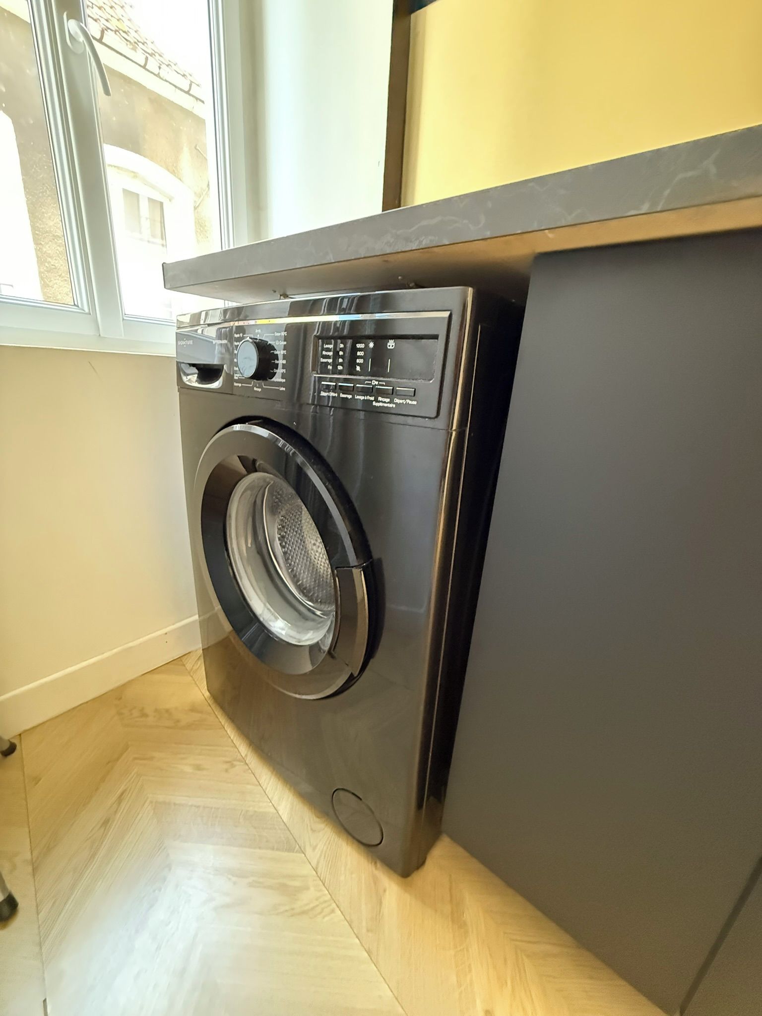 Un lave-linge à chargement frontal se trouve dans une pièce dotée d’une fenêtre et d’un plancher en bois.