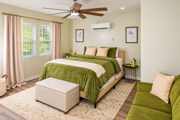 A bedroom with a bed, a ceiling fan, an air conditioning unit, a bedside table, a nightstand, a window with curtains, and a green couch. - Exclusive Queen Suite: Louisville City Escape! (Clarksville)