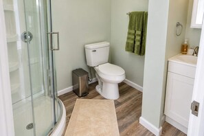 A bathroom with a glass shower enclosure, a toilet, a trash bin, and a green towel hanging on the wall. - Exclusive Queen Suite: Louisville City Escape! (Clarksville)