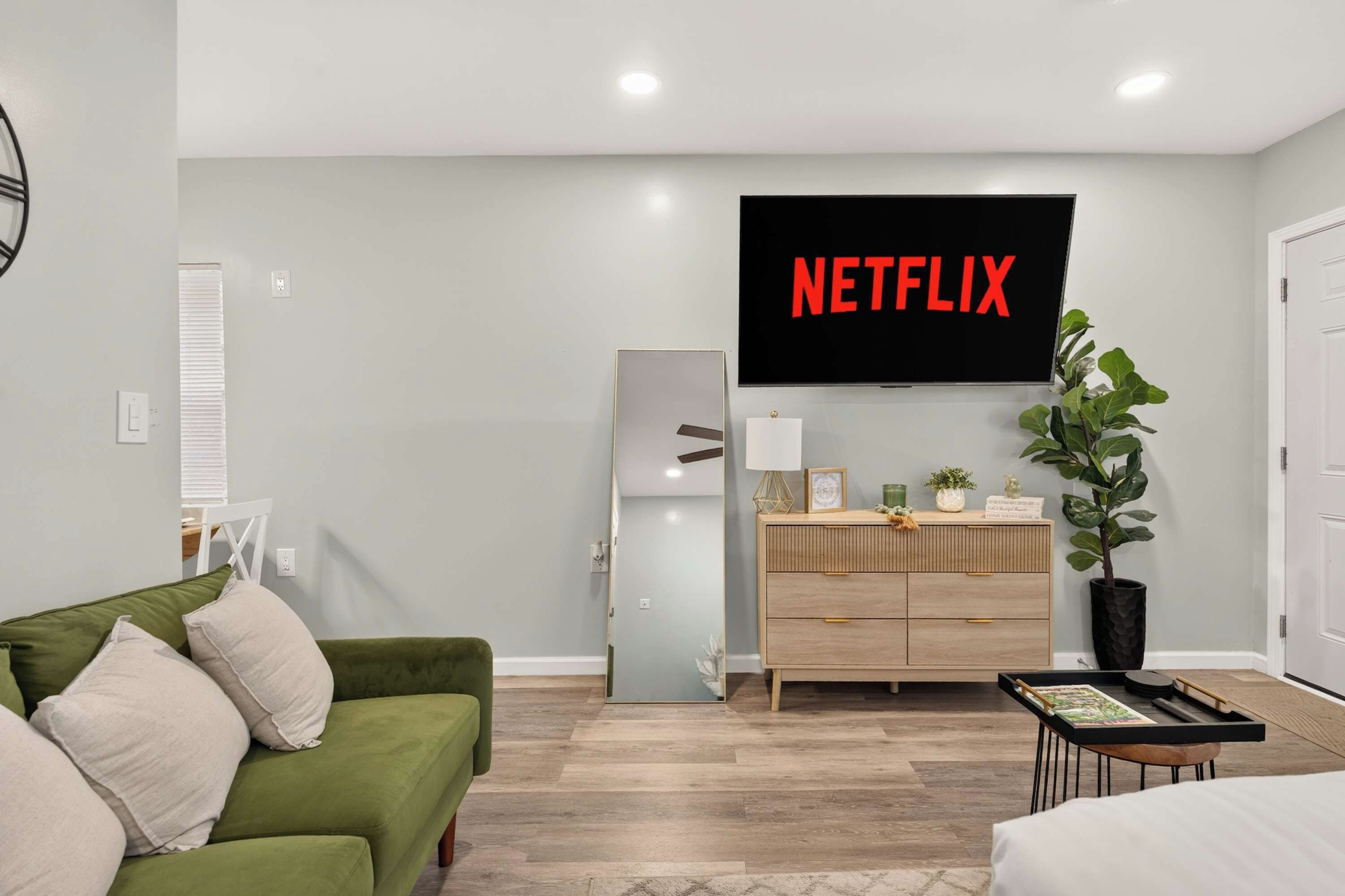 A modern living room with a green sofa, a wooden cabinet, a flat-screen TV mounted on the wall, and a potted plant.