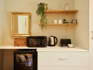 A kitchenette with a microwave, toaster, kettle, and a small refrigerator. There are wooden shelves above holding a plant, a glass, and some kitchen items. A mirror with a gold frame is on the wall.