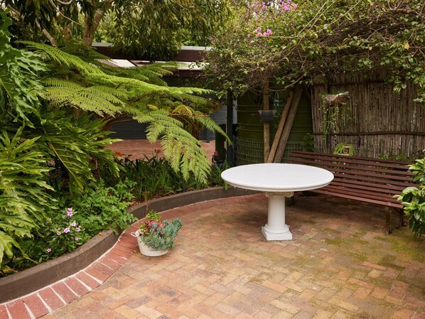 A patio with a white table and bench, surrounded by lush greenery and a brick border.