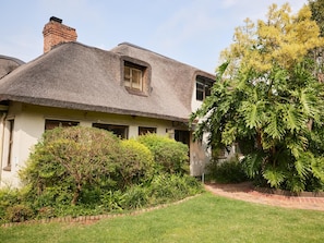 A house with a thatched roof, a visible chimney, and a well-maintained garden with various shrubs and a lawn.
