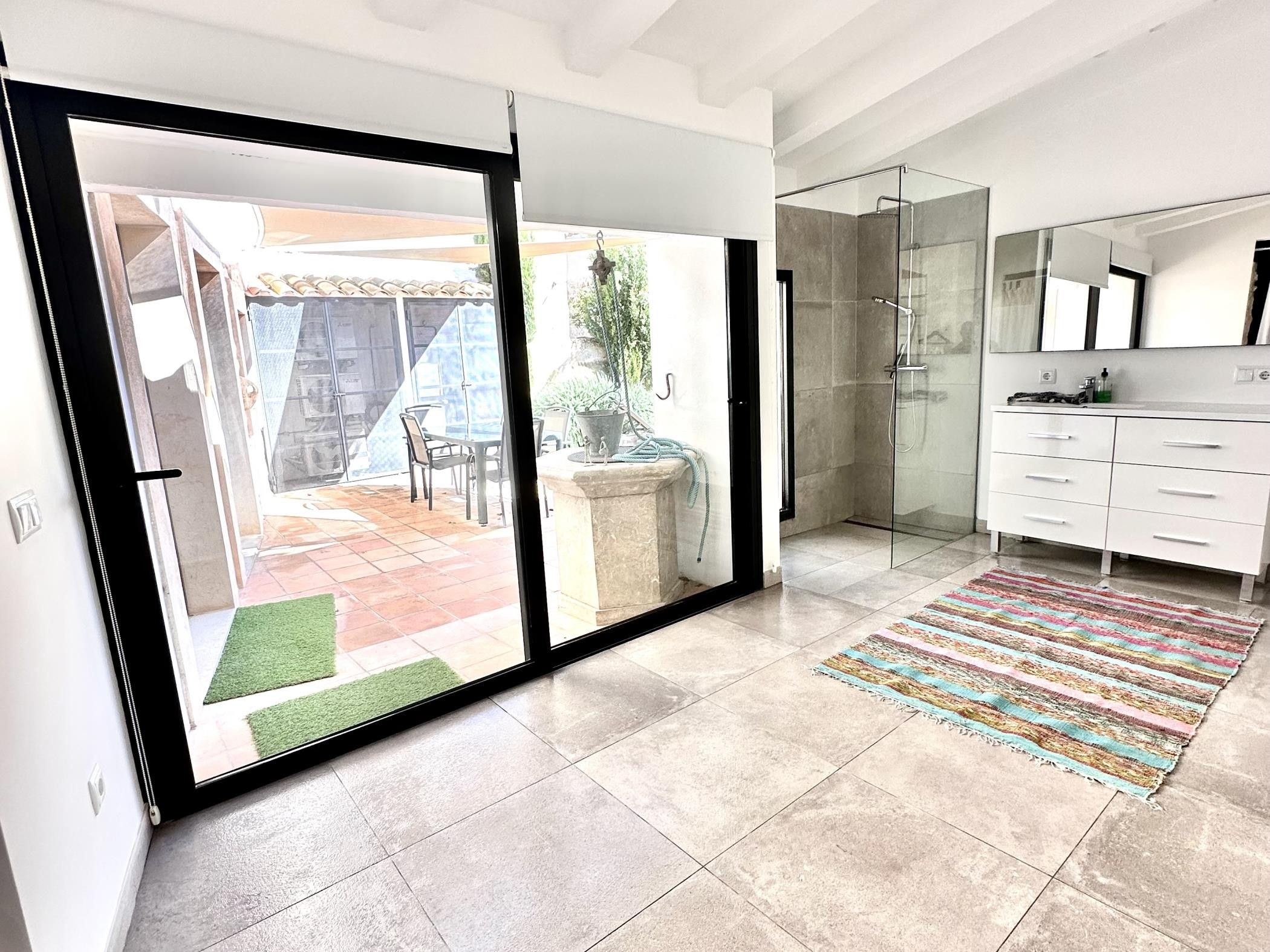 Une salle de bain moderne avec une douche à parois de verre, un meuble-lavabo blanc et une vue sur un patio extérieur doté d’un sol carrelé et d’une plante en pot.