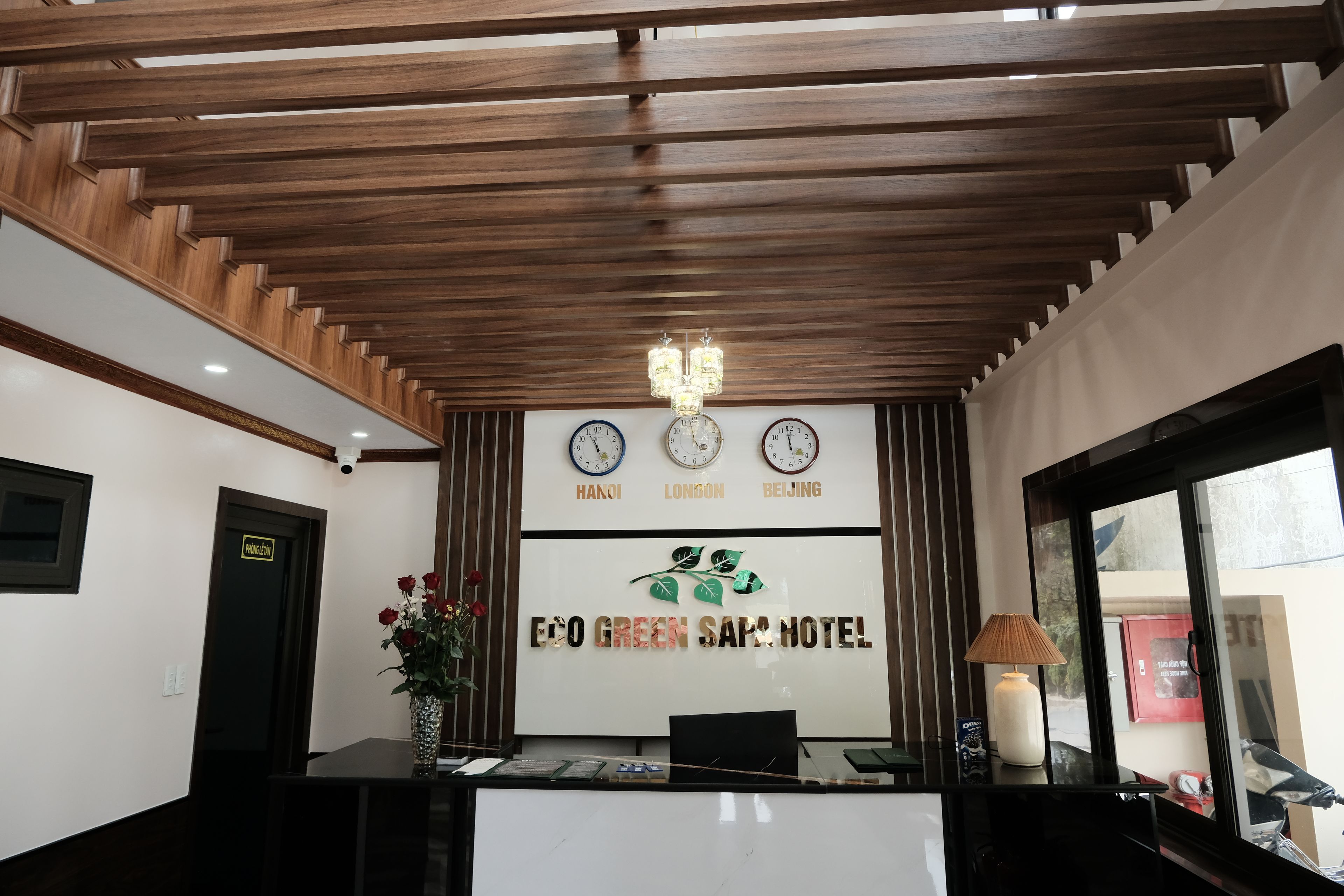 Una recepción con relojes que muestran la hora de Hanói, Londres y Pekín, un cartel que dice EGO GREEN Sapa HOTEL y una cámara de seguridad arriba.