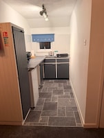 Un pasillo estrecho que conduce a una cocina con un ventanal azul, un lavabo y un hervidor.