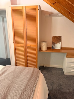 A bedroom with a bed, a wooden wardrobe, a desk with a chair, and a small window.