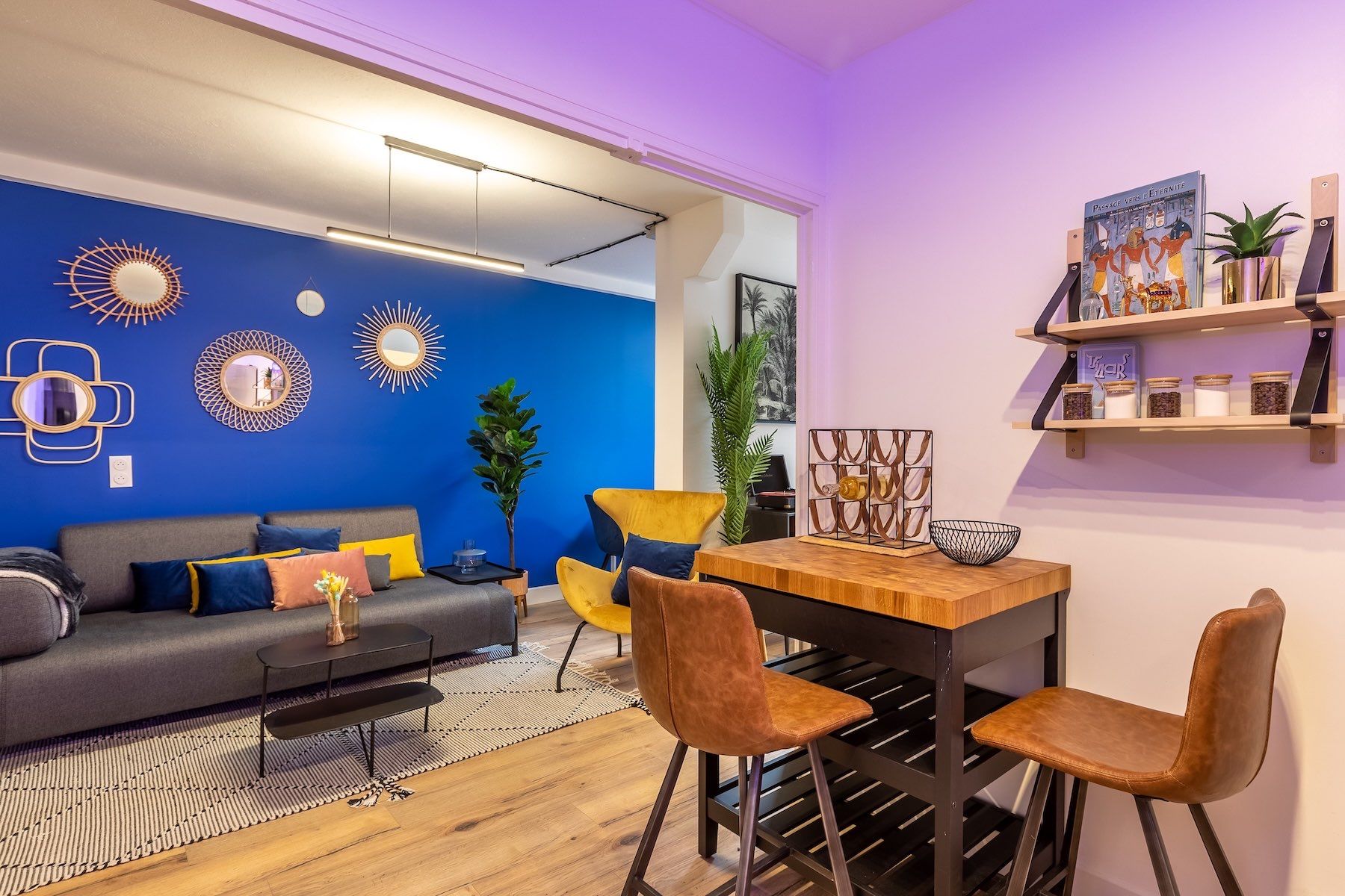 A modern living room with a blue accent wall, a grey sofa, yellow chairs, and a wooden dining table.