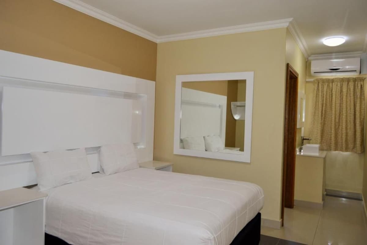 A hotel room with a bed, a bedside table, a mirror, an air conditioning unit, and a bathroom with a sink and toilet.