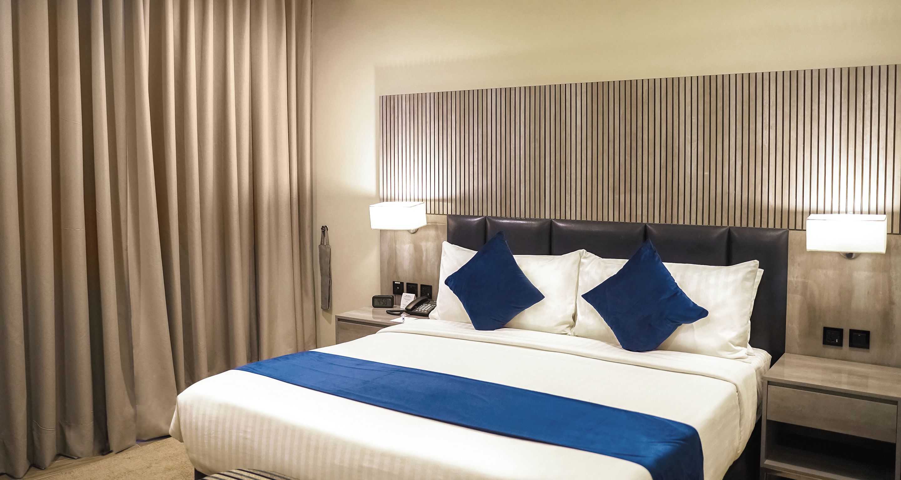 Une chambre d’hôtel avec un lit, des tables de chevet, une petite table, une lampe et un rideau.