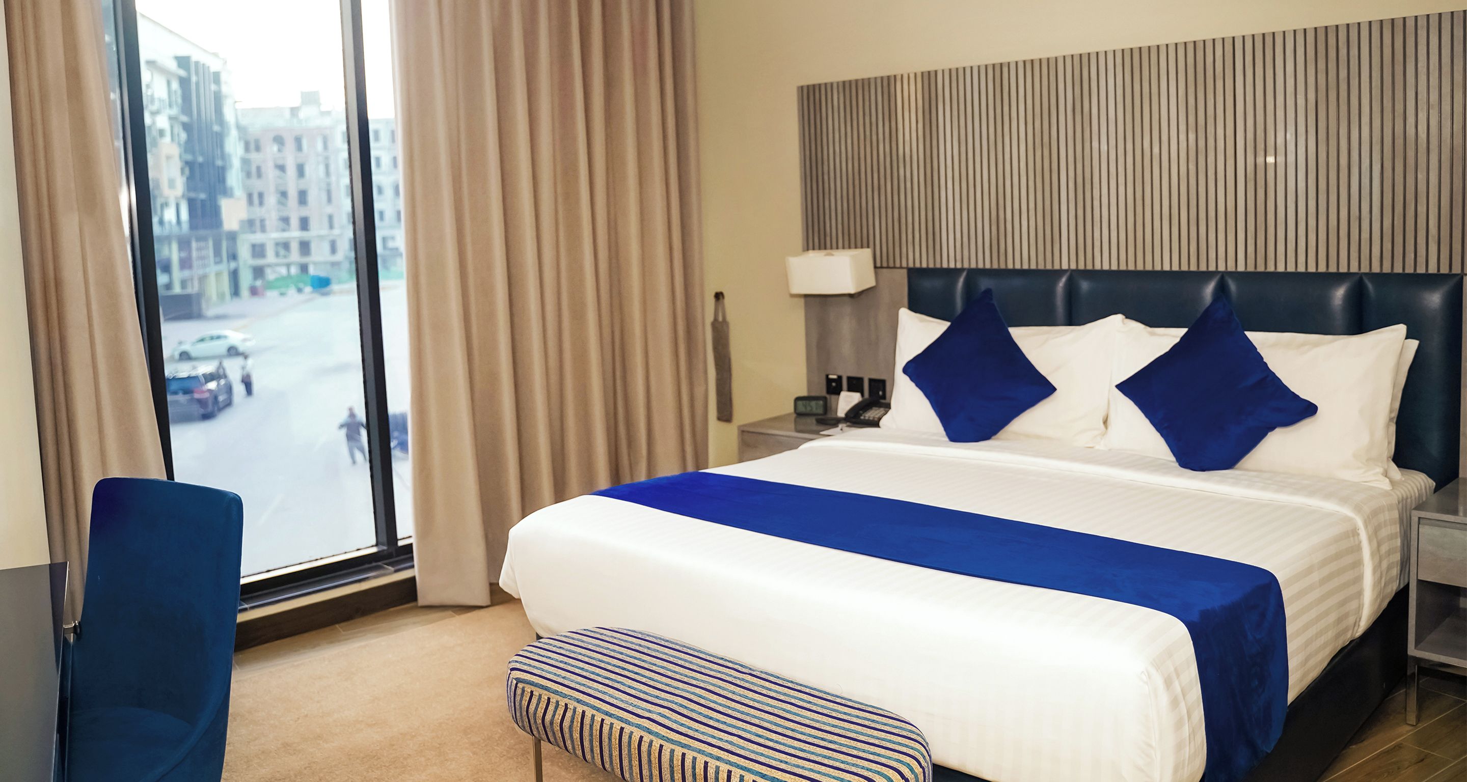 A hotel room with a large bed, a bedside table with a lamp, a chair, and a view of the street through a window.