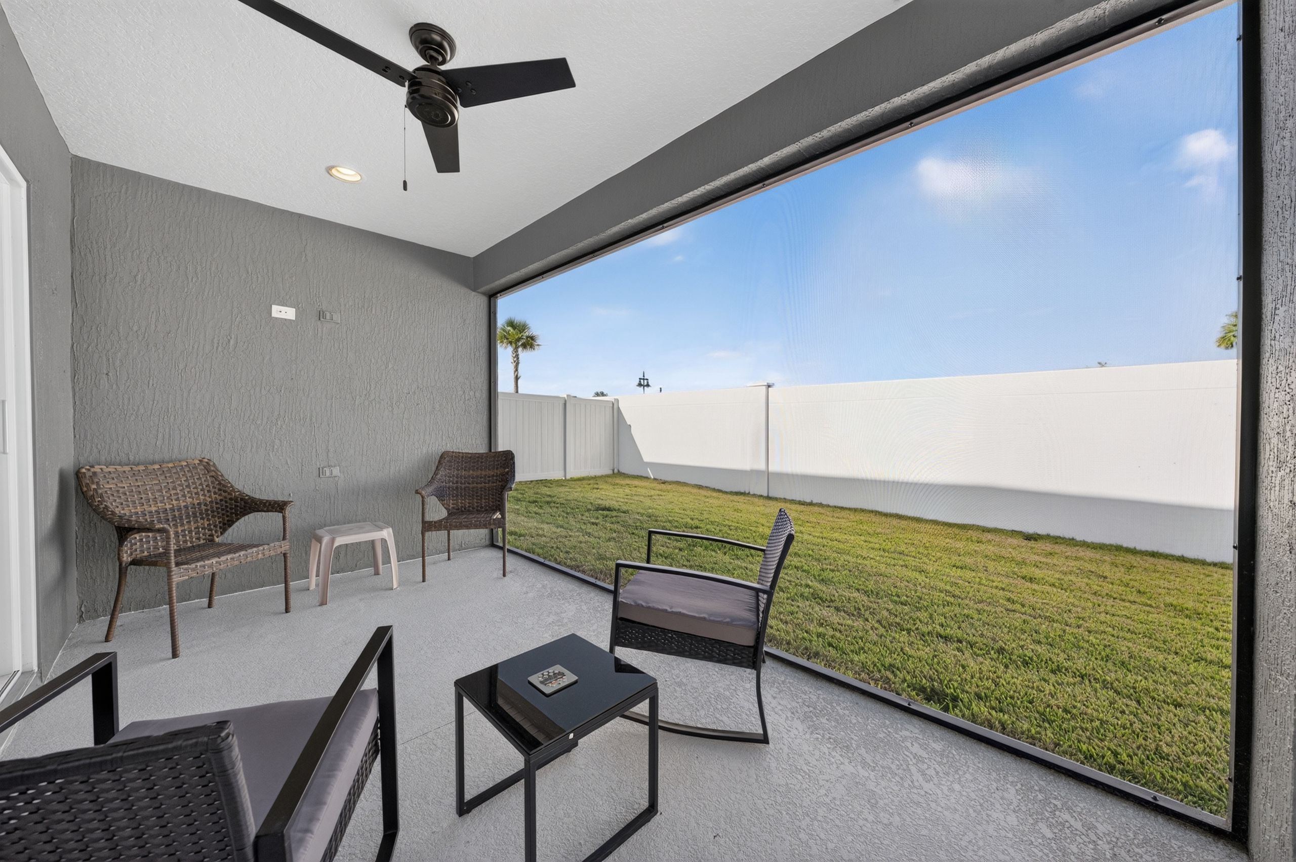 A modern outdoor patio with a ceiling fan, two wicker chairs, a small table, and a view of a grassy area.