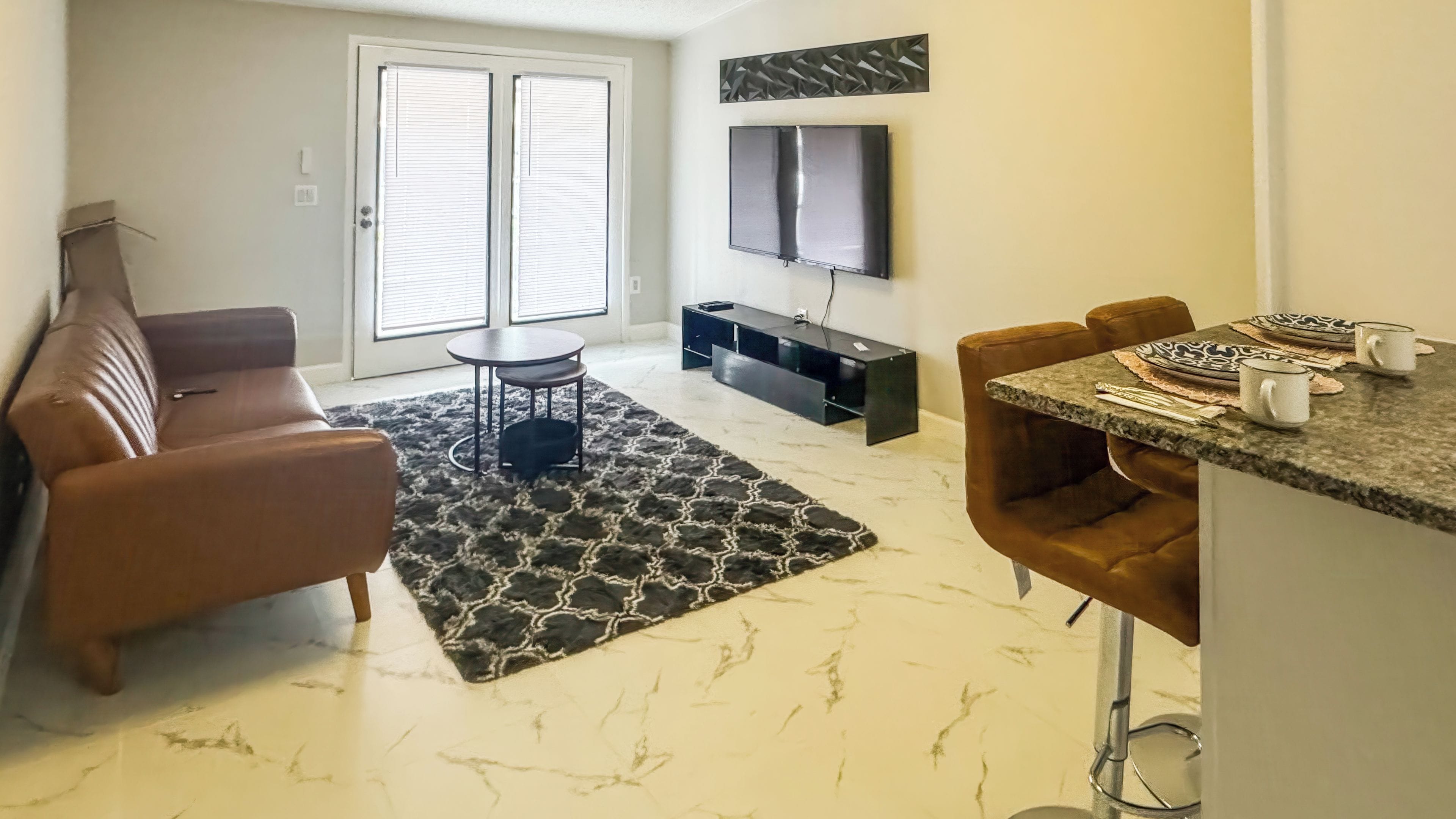 A living room with a brown leather sofa, a round coffee table, a black rug, a TV mounted on the wall, and a dining area with a granite countertop and bar stools.