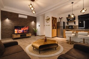 A modern living room with a flat-screen TV, a wooden coffee table, and a kitchen area with bar stools. - New Listing (Yaoundé)