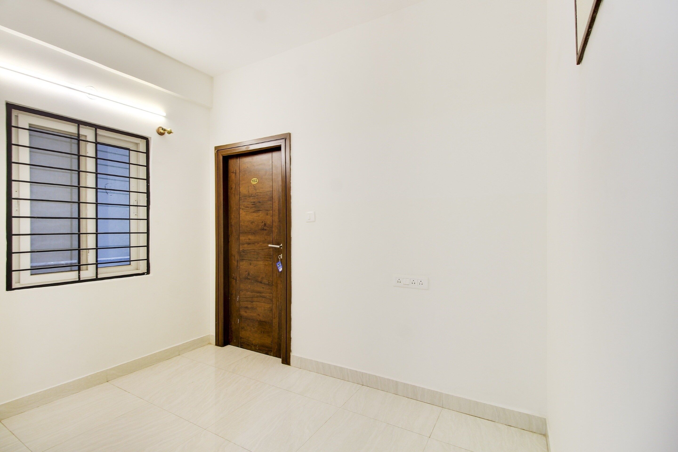A hallway with a wooden door marked with the number 102, a window with metal bars, and a light switch on the wall.