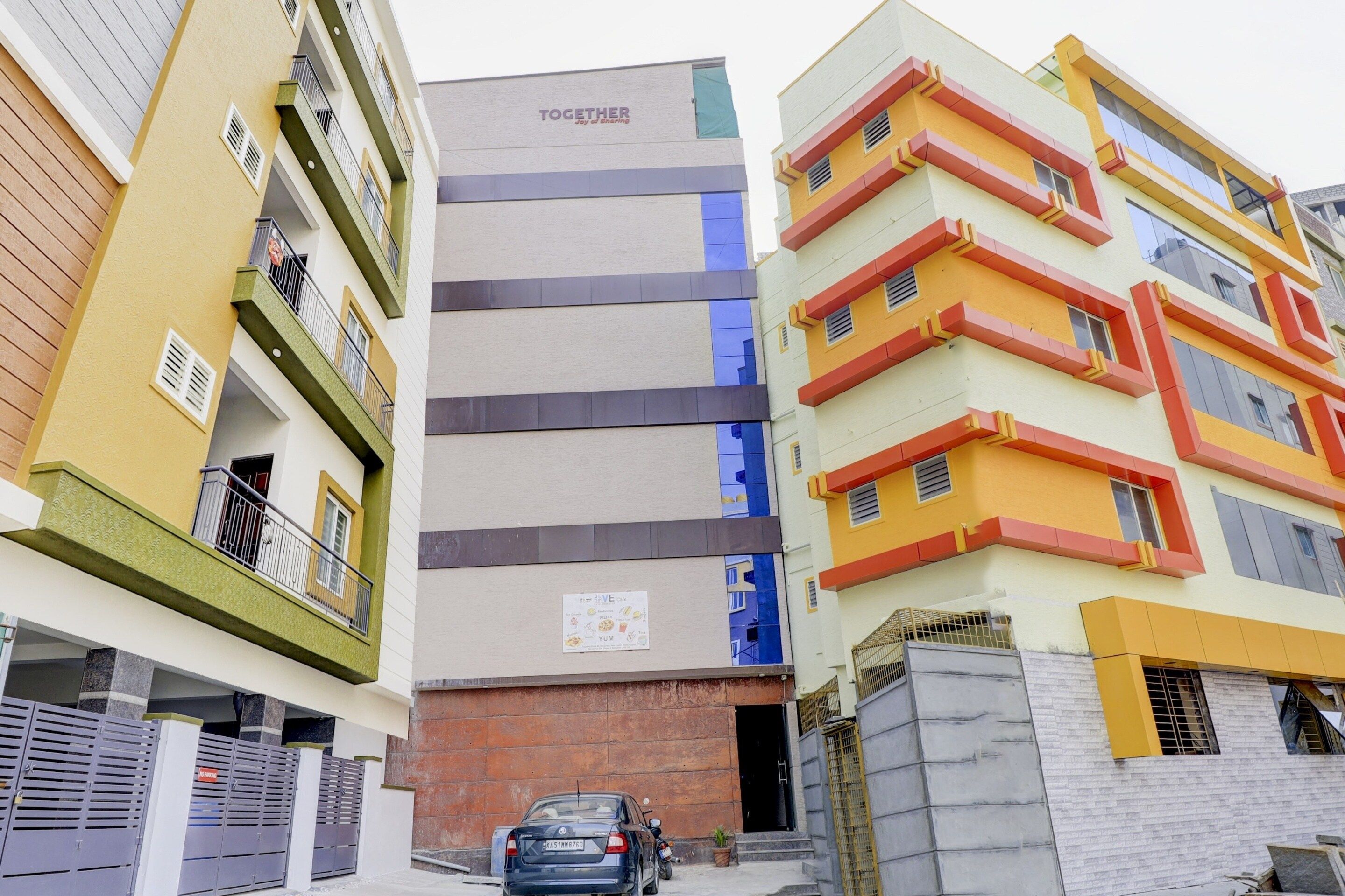 Un edificio residencial de varios pisos con balcones, un letrero visible de una cafetería y un coche estacionado en la calle.