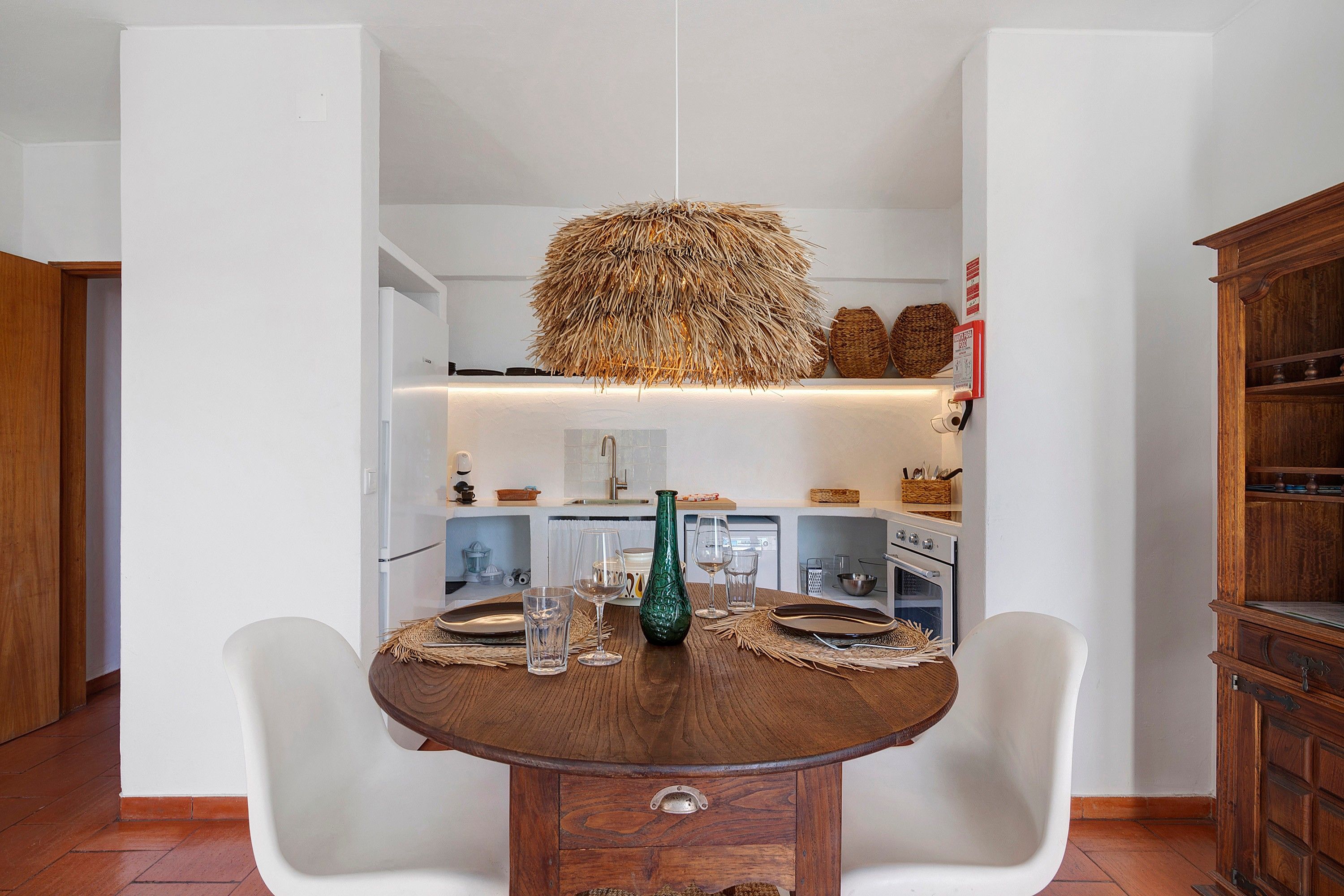 Un coin repas avec une table ronde en bois, des chaises blanches, un luminaire suspendu et une cuisine en arrière-plan.