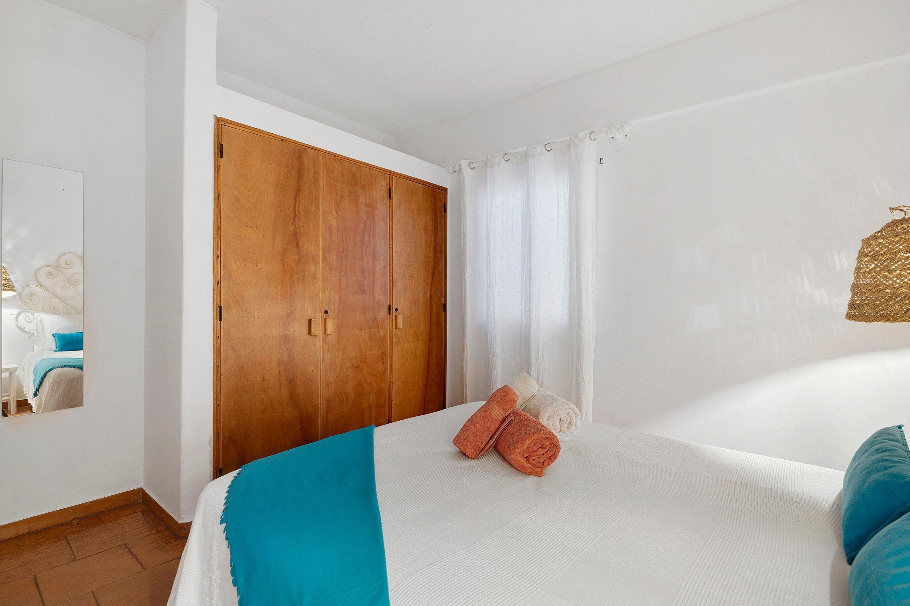 Une chambre à coucher avec un lit, une armoire en bois, un rideau blanc et un luminaire fixé au mur.