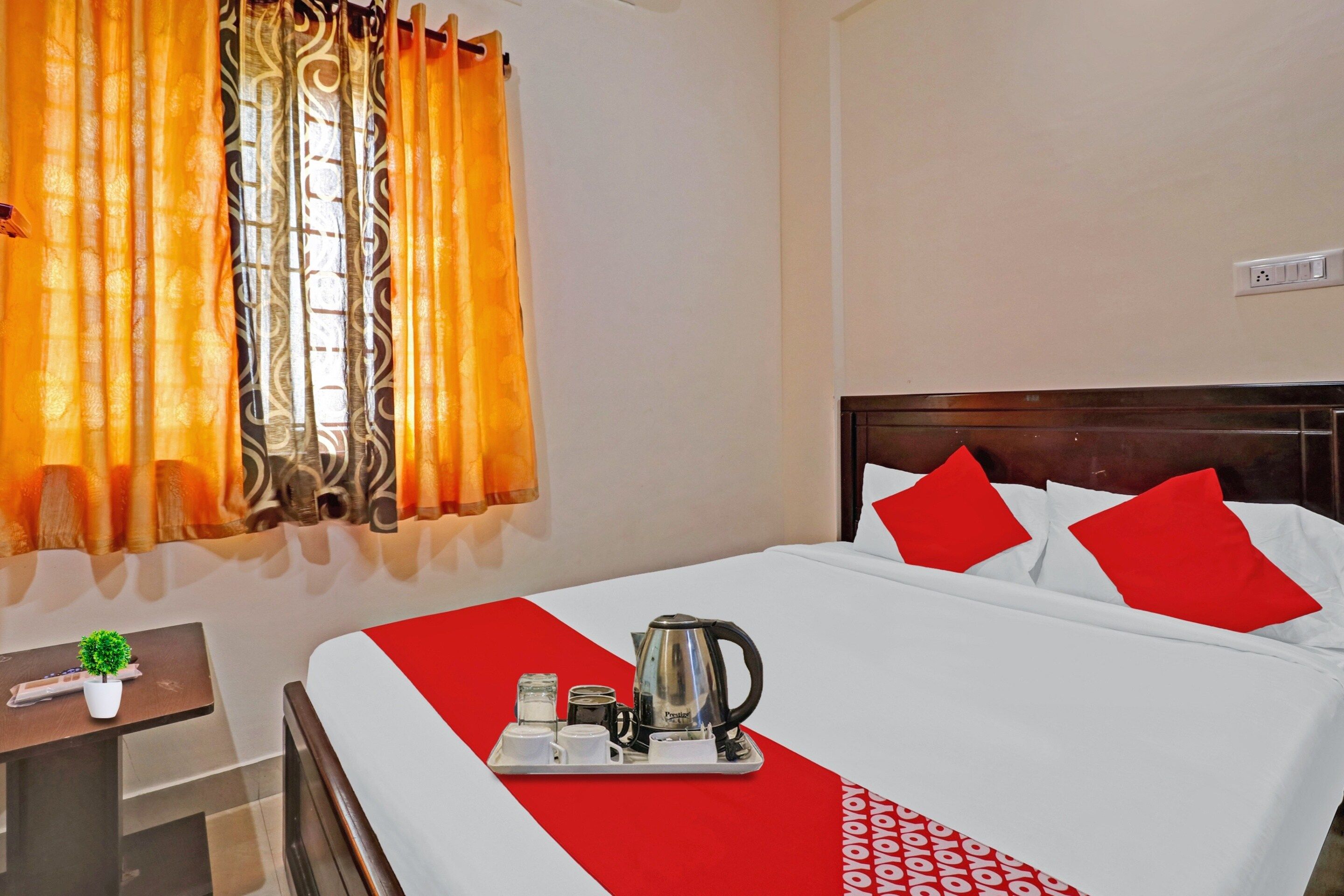 A hotel room with a bed, a nightstand, a window with curtains, and a tray with a kettle and cups.
