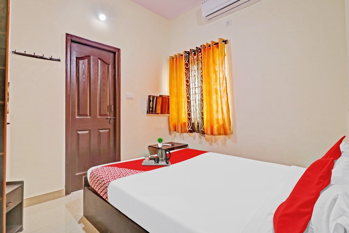 A hotel room with a bed, a bedside table with a kettle, a small plant, and a window with curtains.