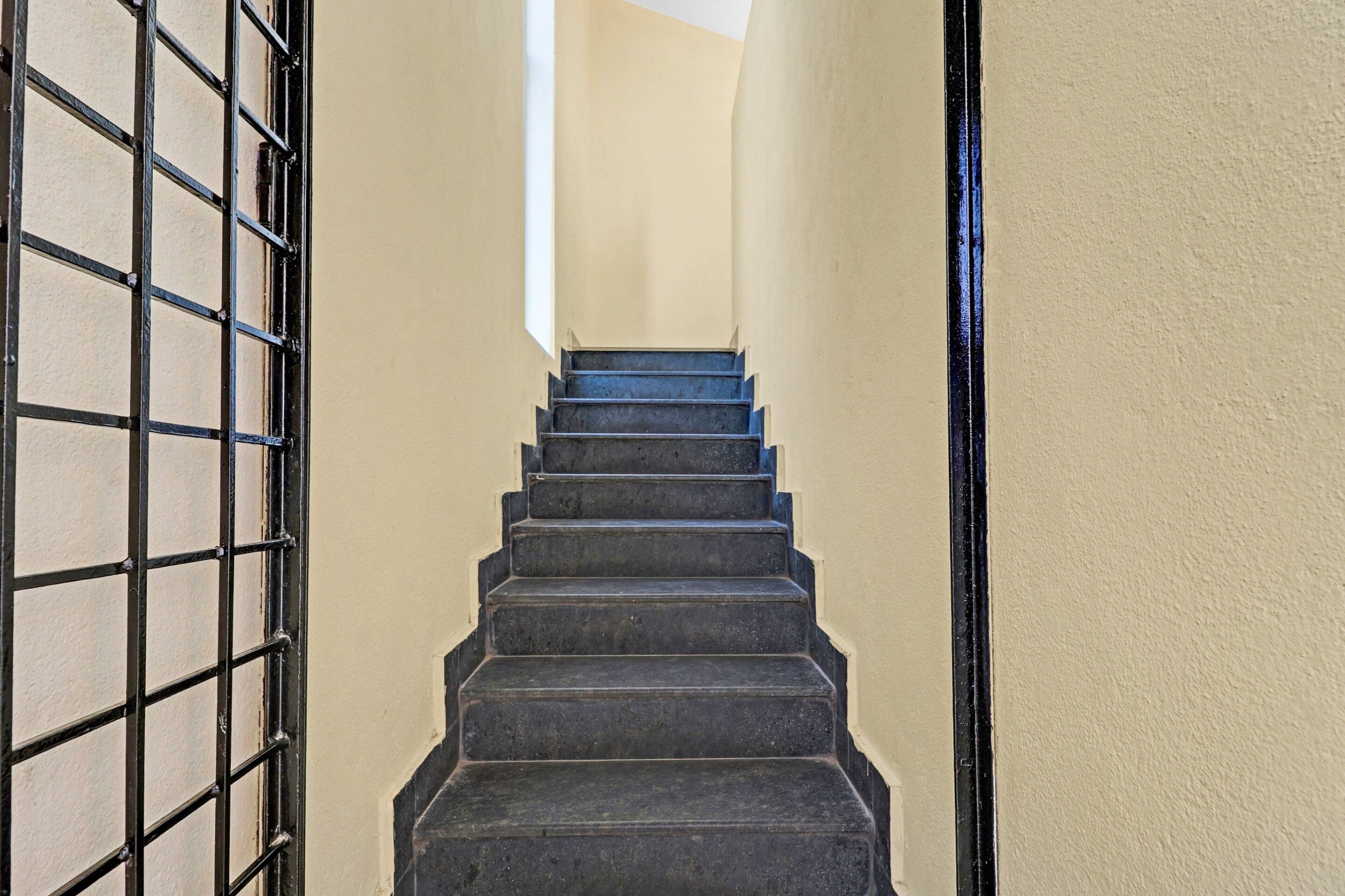 A staircase with black steps leading up to a landing, flanked by beige walls and a metal gate on the left.