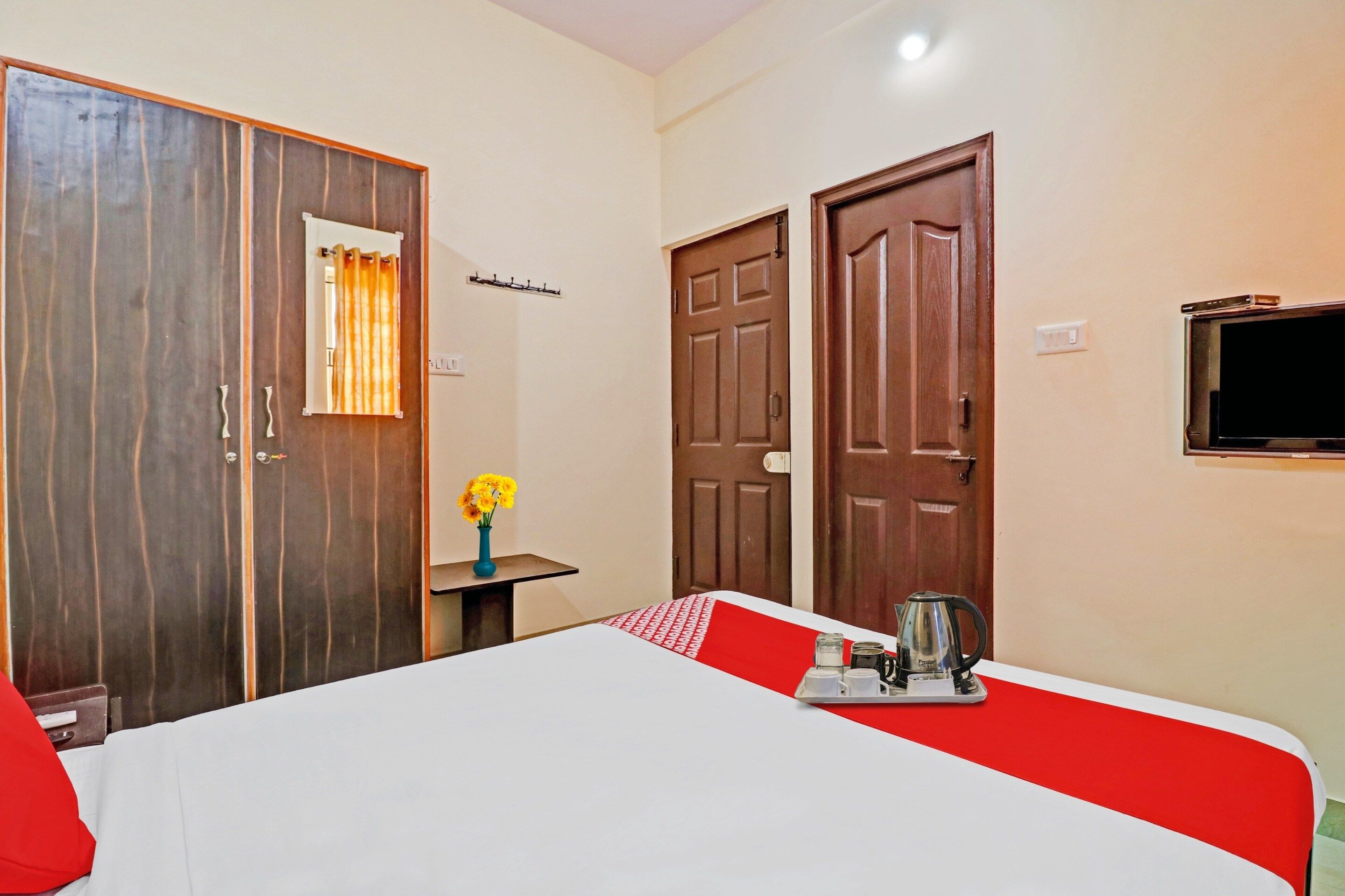 A hotel room with a bed, a small table with a kettle and cups, a vase with flowers, and two doors.
