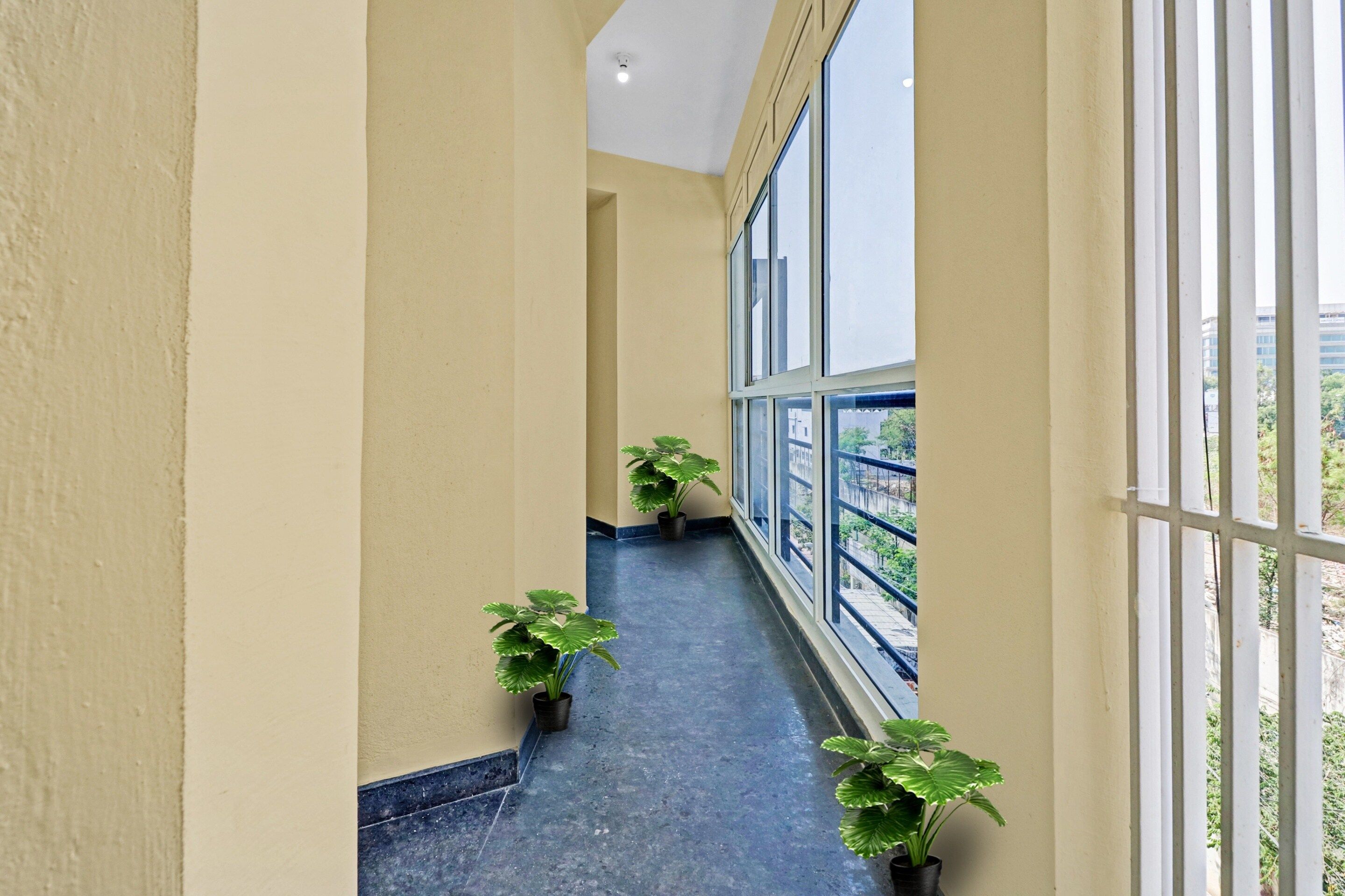 A corridor with a blue floor, yellow walls, and large windows on one side. There are potted plants placed along the corridor.