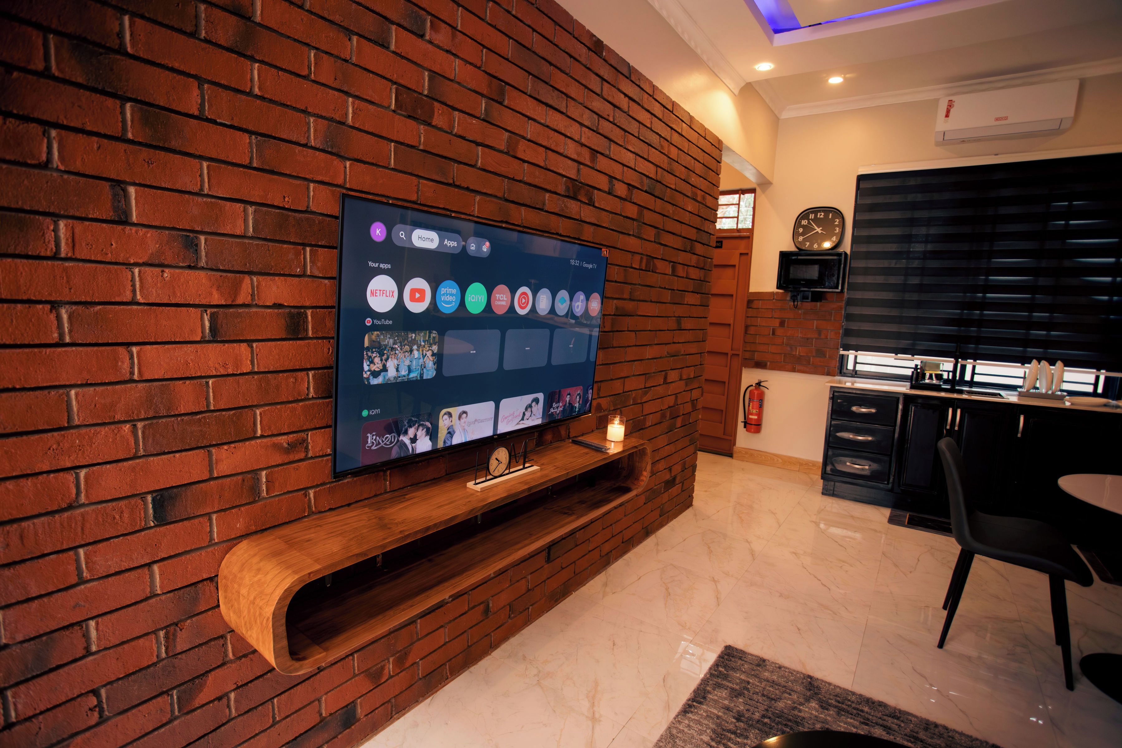 Ein modernes Wohnzimmer mit Flachbildfernseher an einer Ziegelwand, einer Holz-Unterhaltungsecke darunter und einem Essbereich mit Tisch und Stühlen.