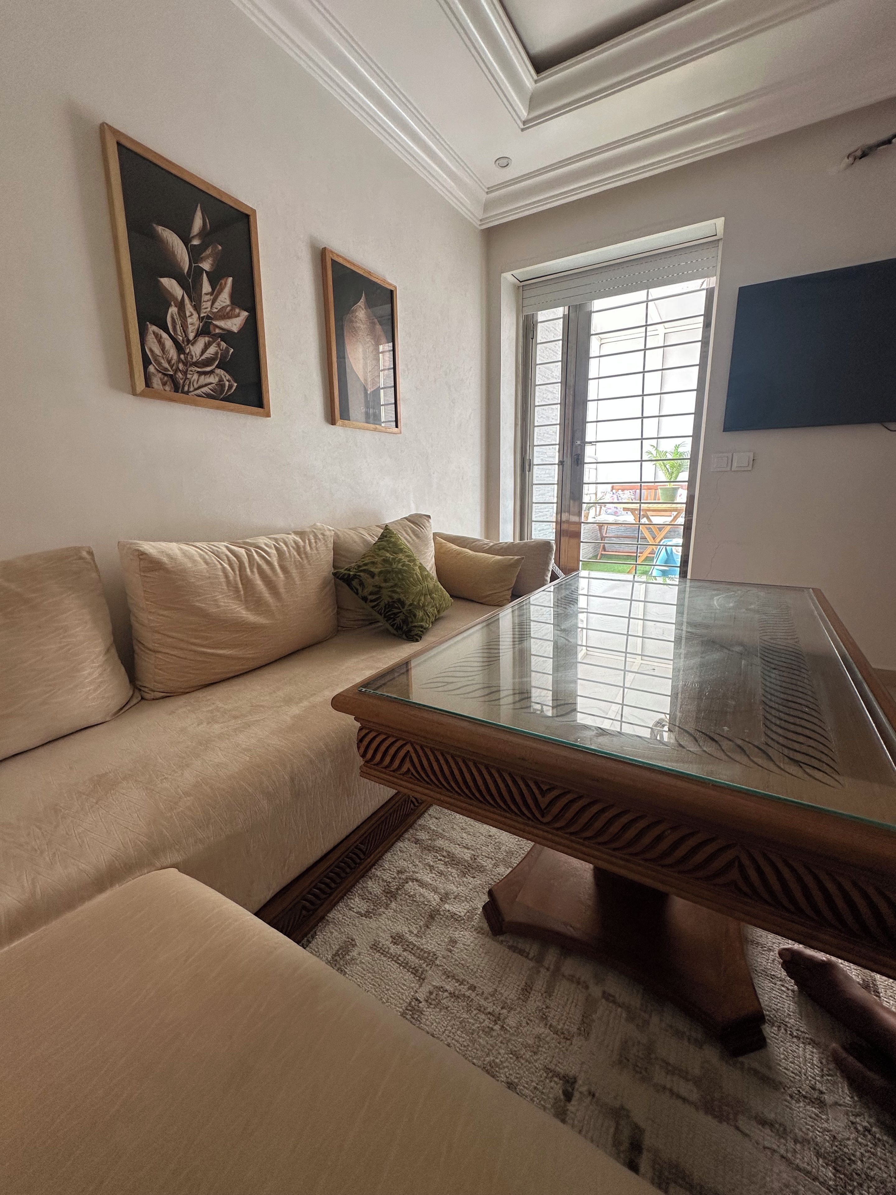 Un salon avec une table basse en verre, un canapé beige et deux œuvres d’art encadrées accrochées au mur.