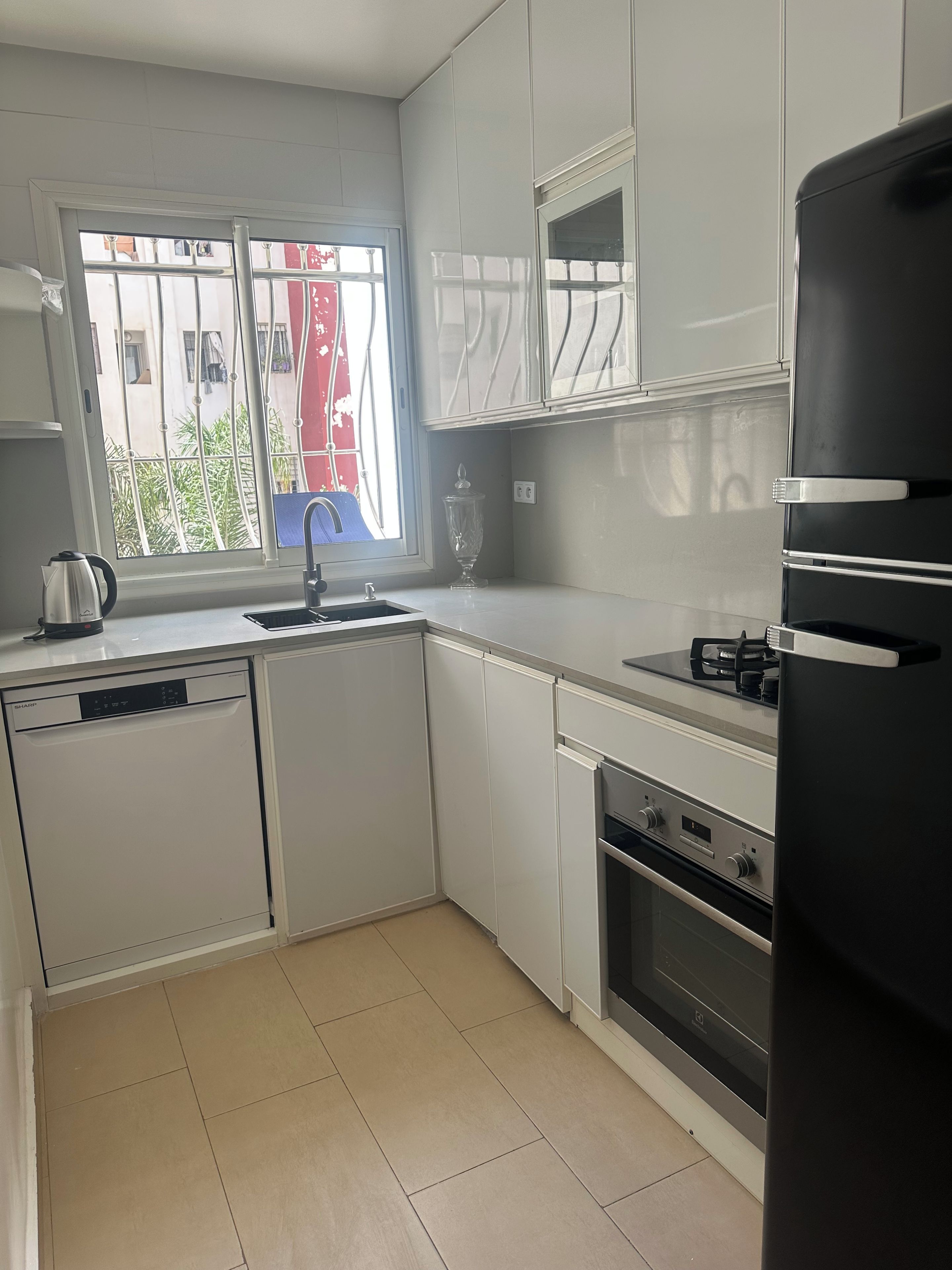 Une cuisine moderne avec des meubles blancs, un four encastré, un lave-vaisselle et une fenêtre donnant sur un bâtiment rouge et sur la verdure environnante.