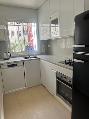Una cuina moderna amb armaris blancs, forn integrat, rentaplats i finestra amb vista a un edifici vermell i vegetació exterior.
