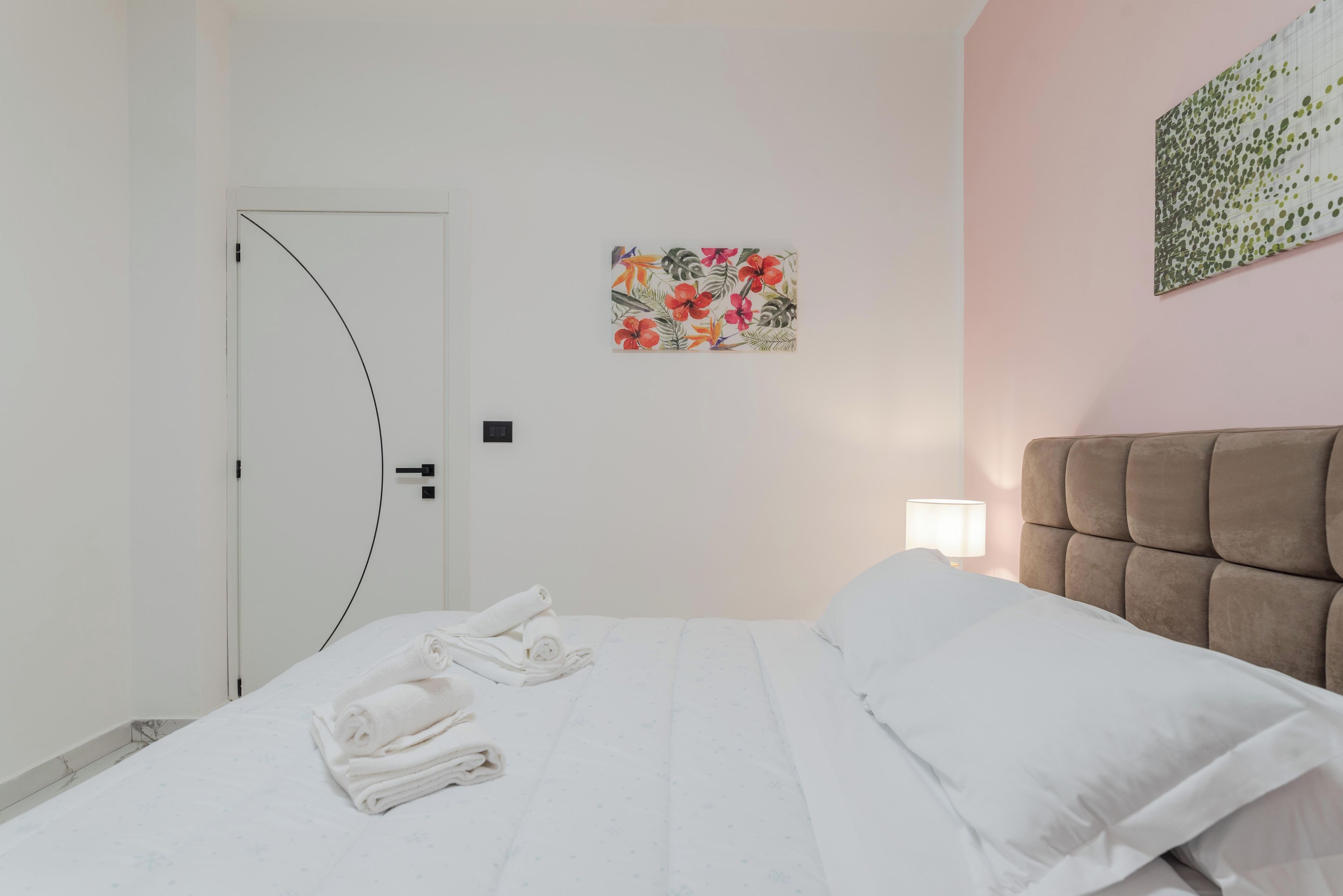 A hotel room with a bed, a door, a wall-mounted artwork, and a bedside lamp.