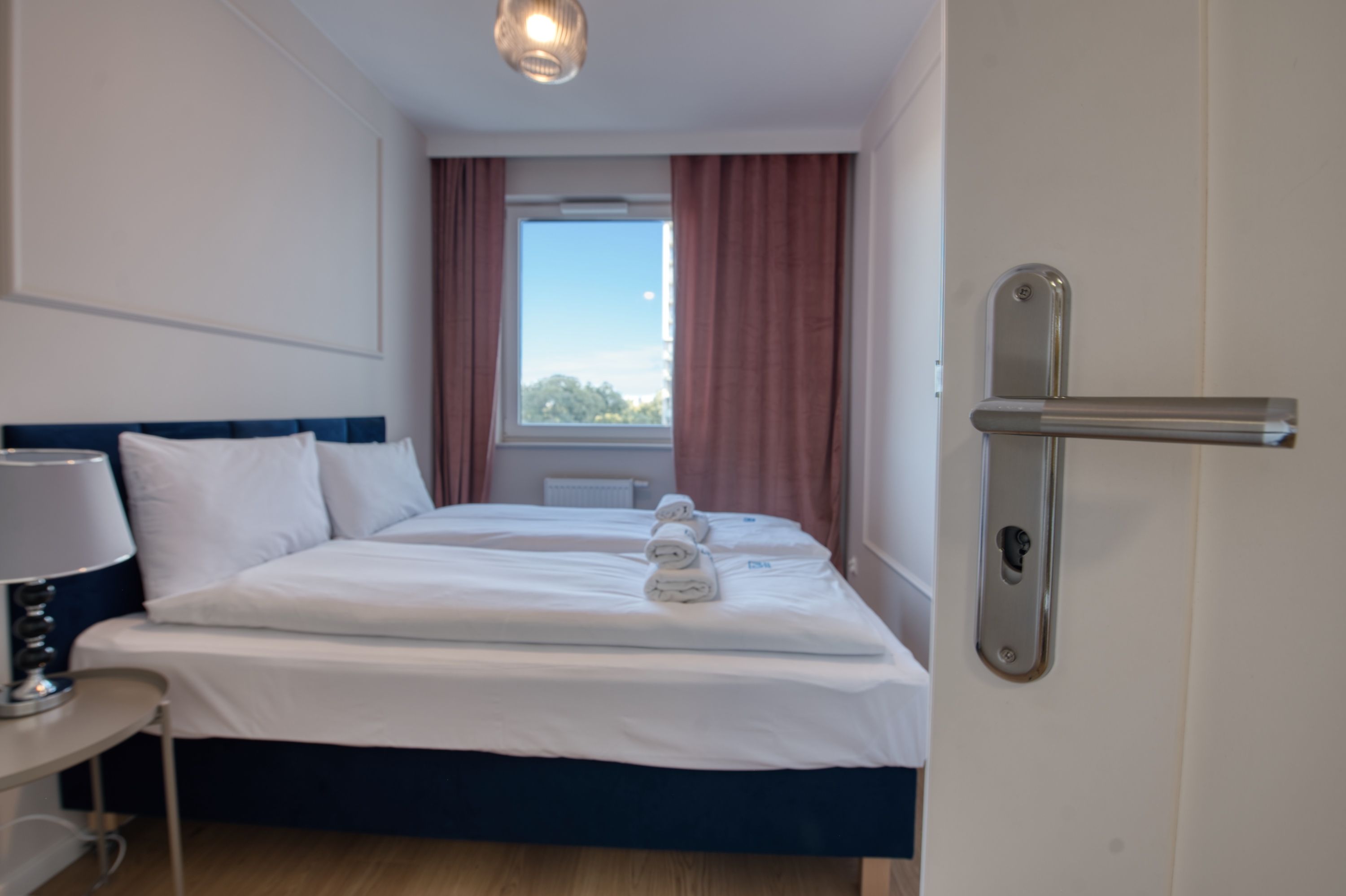 Una habitación de hotel con una cama, una mesita de noche con lámpara, una ventana con cortinas y una puerta con picaporte.