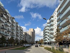 Uma rua urbana moderna com edifícios residenciais altos, céu claro e carros estacionados.