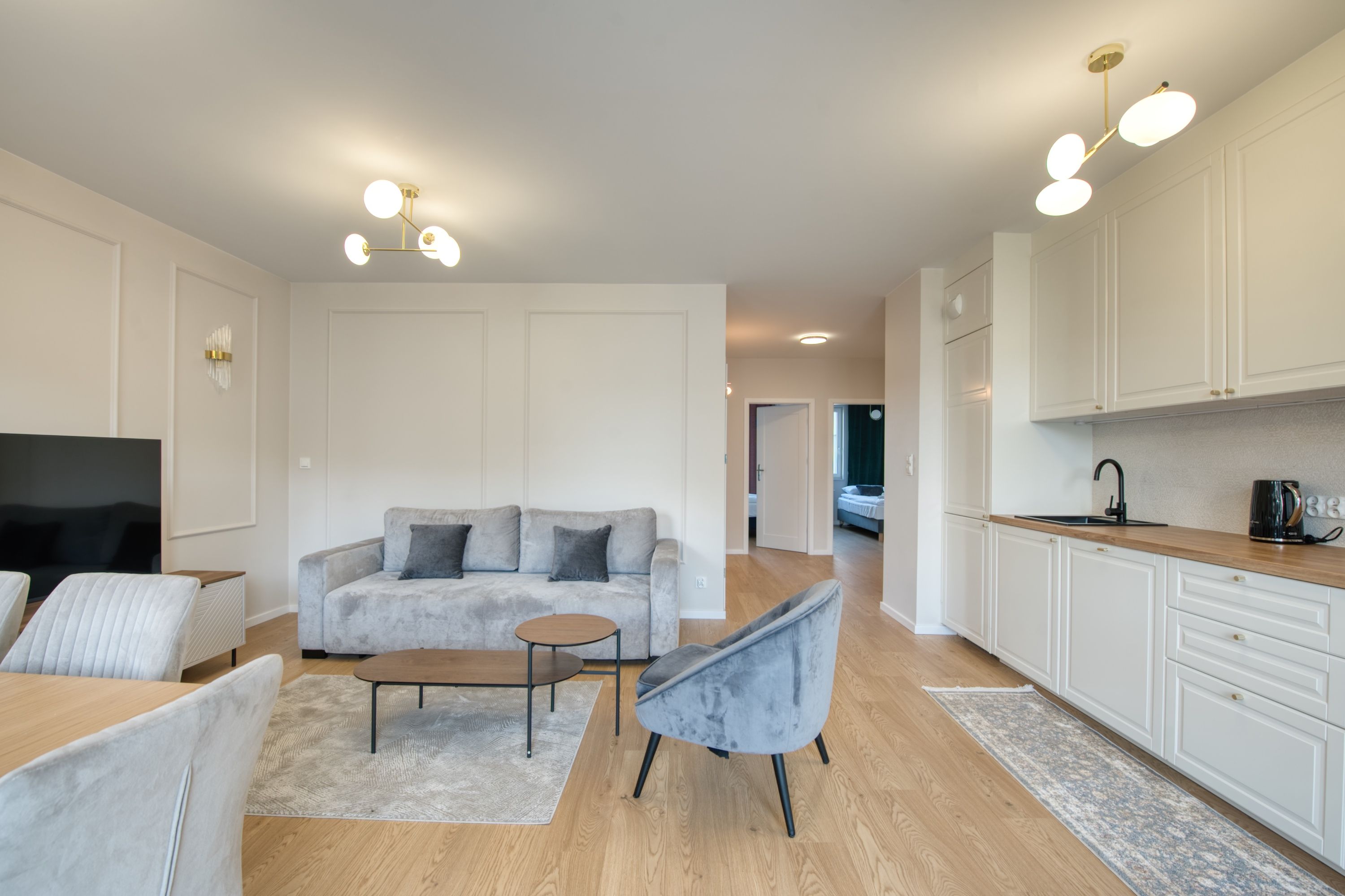 Una sala moderna con un sofá gris, una mesa de centro y un televisor. Se observa una cocina con armarios blancos y un hervidor negro sobre la encimera.