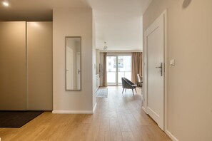 Corredor com piso de madeira, espelho, porta e vista para uma varanda com cortinas.