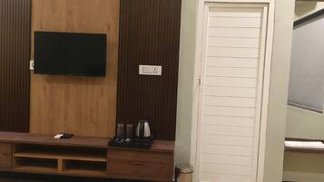 Habitación con un televisor de pantalla plana montado en una pared de madera, una mesita con una tetera, tazas y un control remoto, y una puerta blanca con un espejo.
