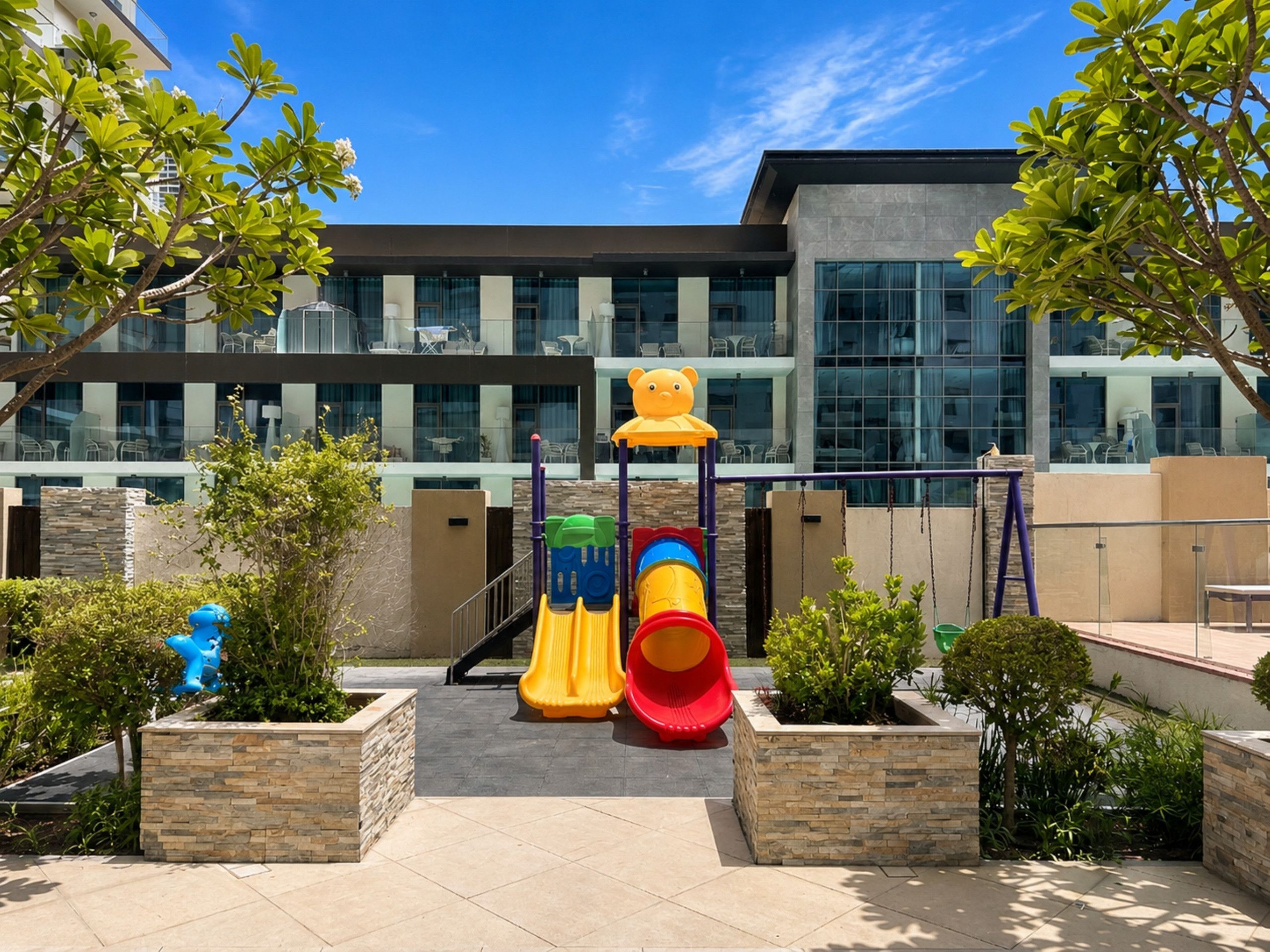 An outdoor play area with slides and climbing structures, surrounded by landscaped gardens and a modern building in the background.