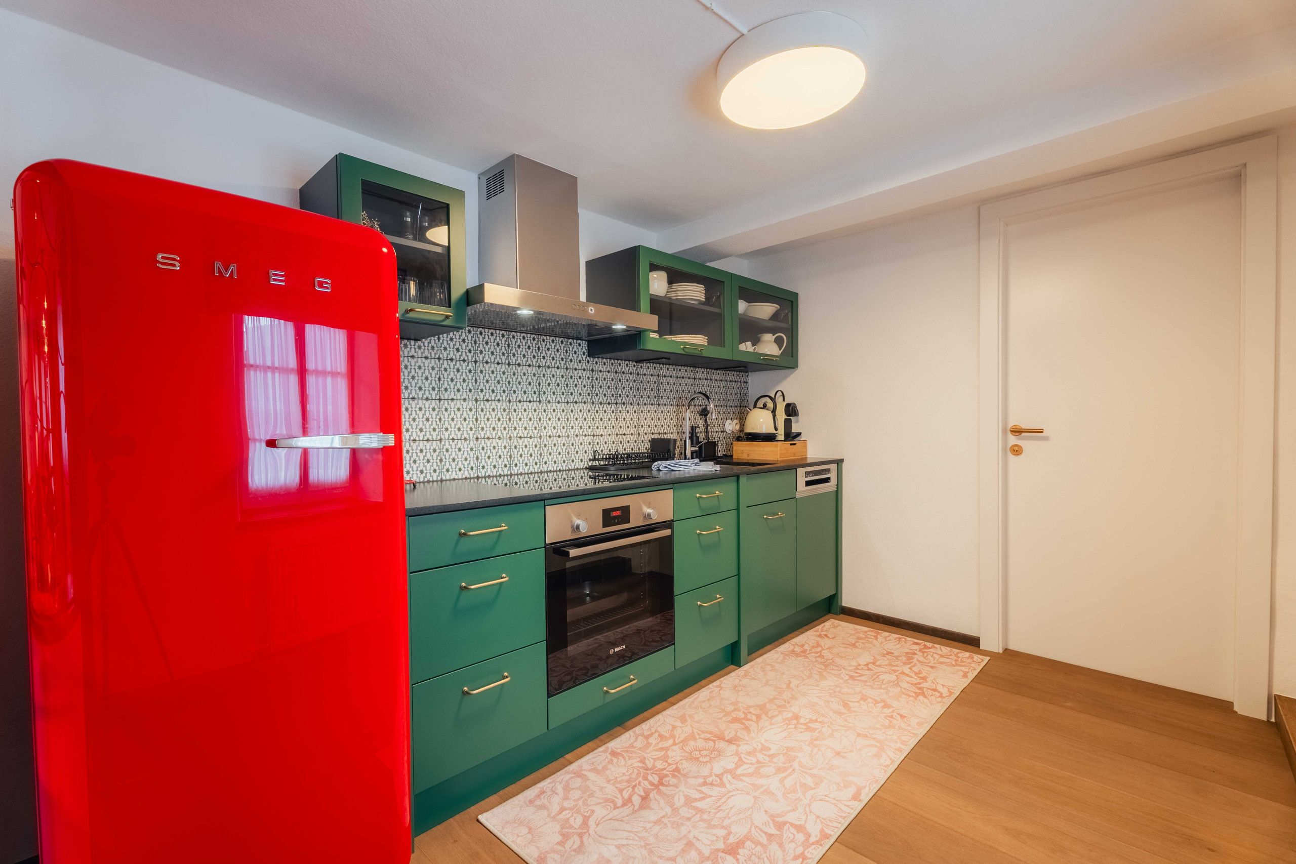Une cuisine de taille modeste, avec un réfrigérateur rouge, des armoires vertes, un pare-feu décoré et un plancher en bois.