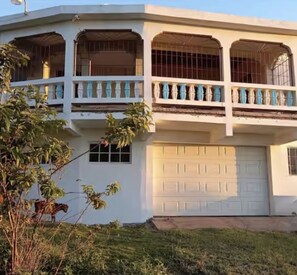 A two-story building with a balcony, a garage, and a small garden area. - Moorish Mount Queen Retreat – Cool Hilltop Room, Mandeville (Mandeville)