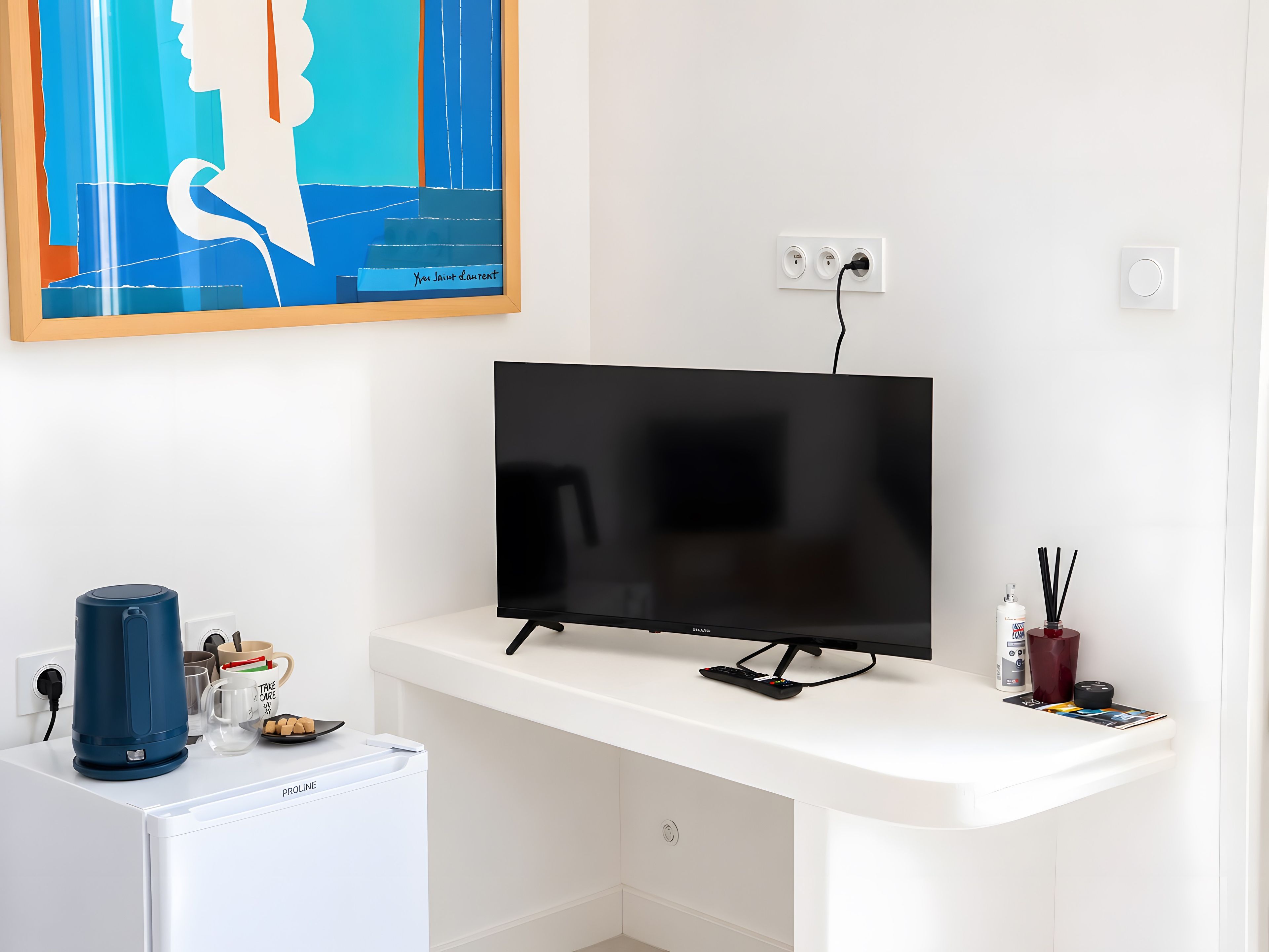 Un salon moderne avec un téléviseur à écran plat posé sur un meuble blanc, une machine à café, un petit réfrigérateur et un tableau accroché au mur.