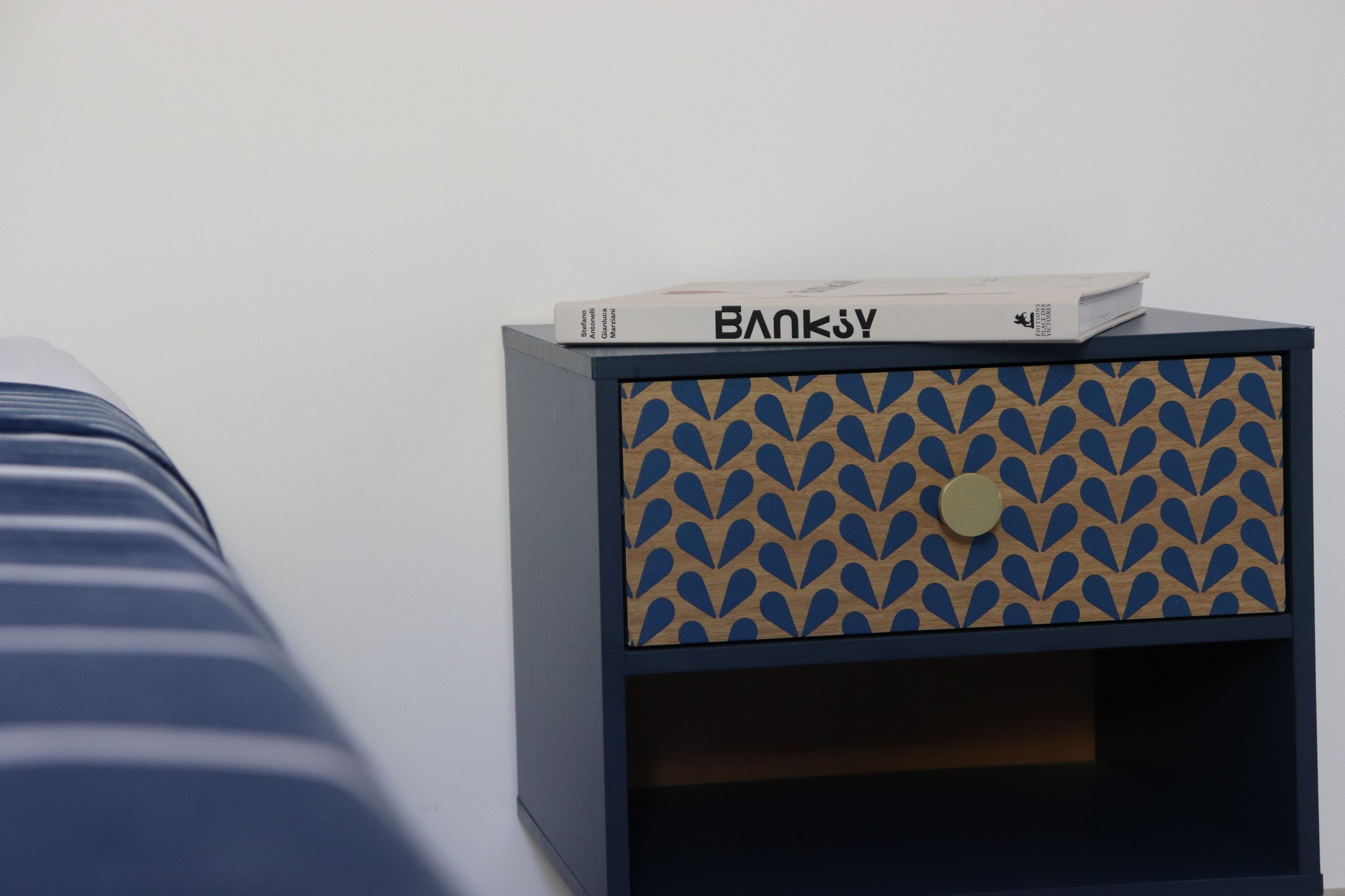 Yöpöytä, jossa on kuvioitu pinta, ja sen päällä on kirja, jonka nimi on ”BANKJY”.