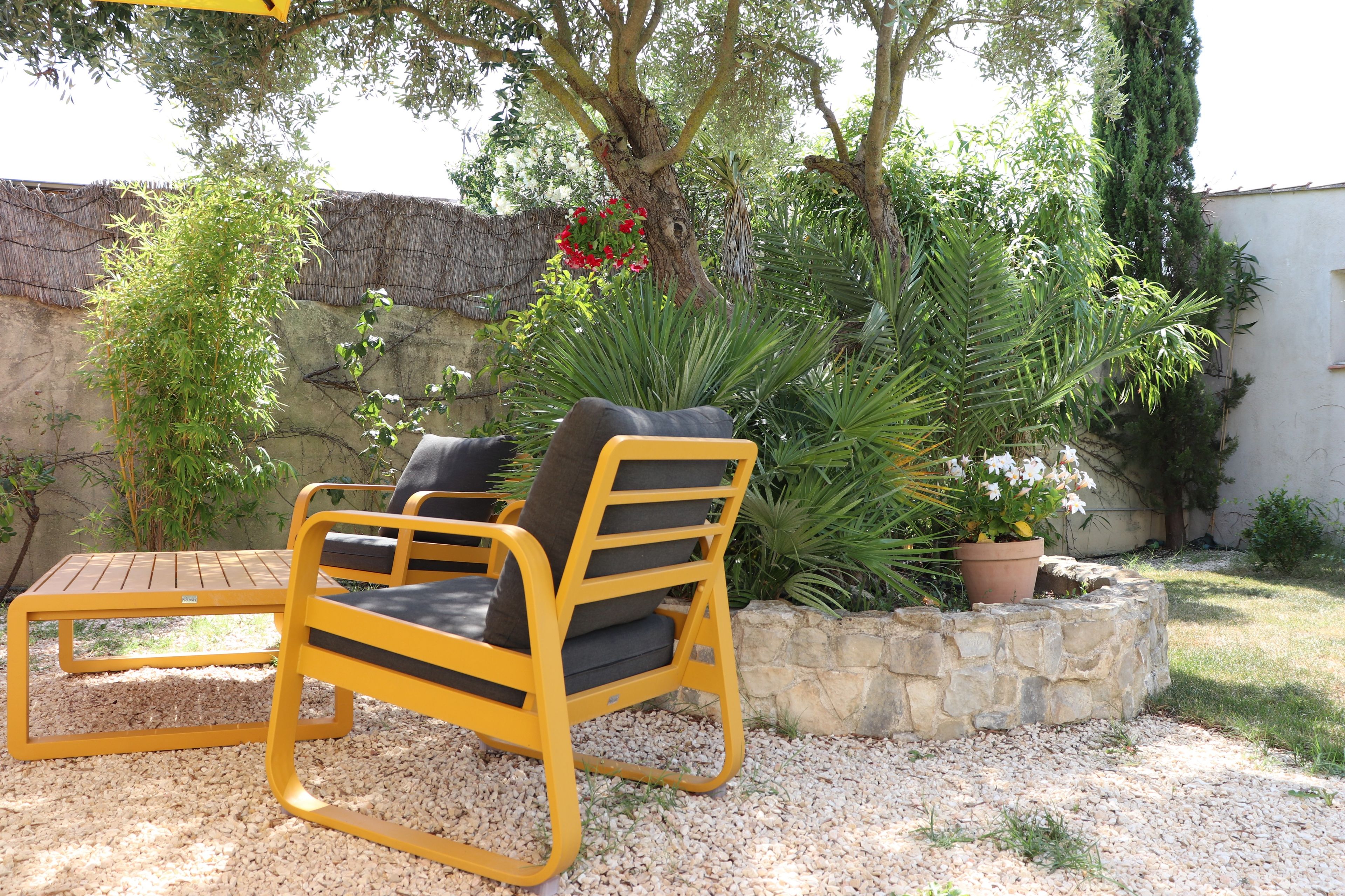 Un área de estar al aire libre con una silla de metal amarillo y una pequeña mesa, rodeada de grava y diversas plantas.