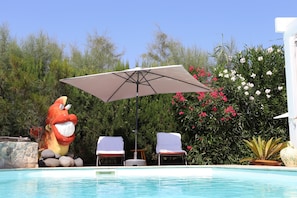 Una piscina con due sdraio e un ombrellone, circondata da vegetazione e una grande statua a forma di personaggio dei cartoni animati.