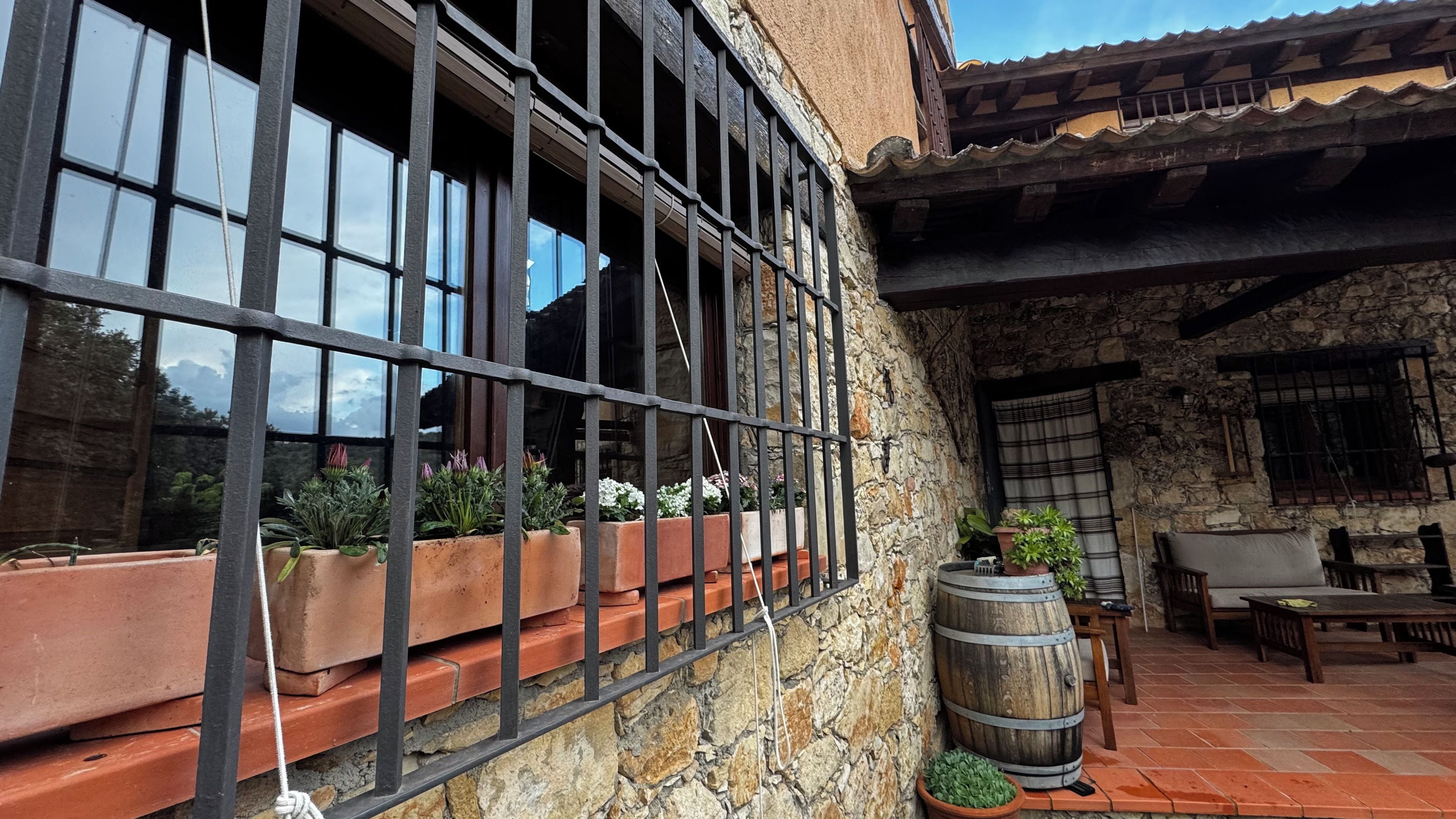 Un edificio de piedra con una ventana de barrotes, macetas, un barril de madera y mobiliario exterior.