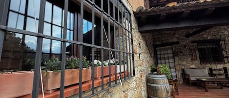 Un edificio de piedra con una ventana de barrotes, macetas, un barril de madera y mobiliario exterior.