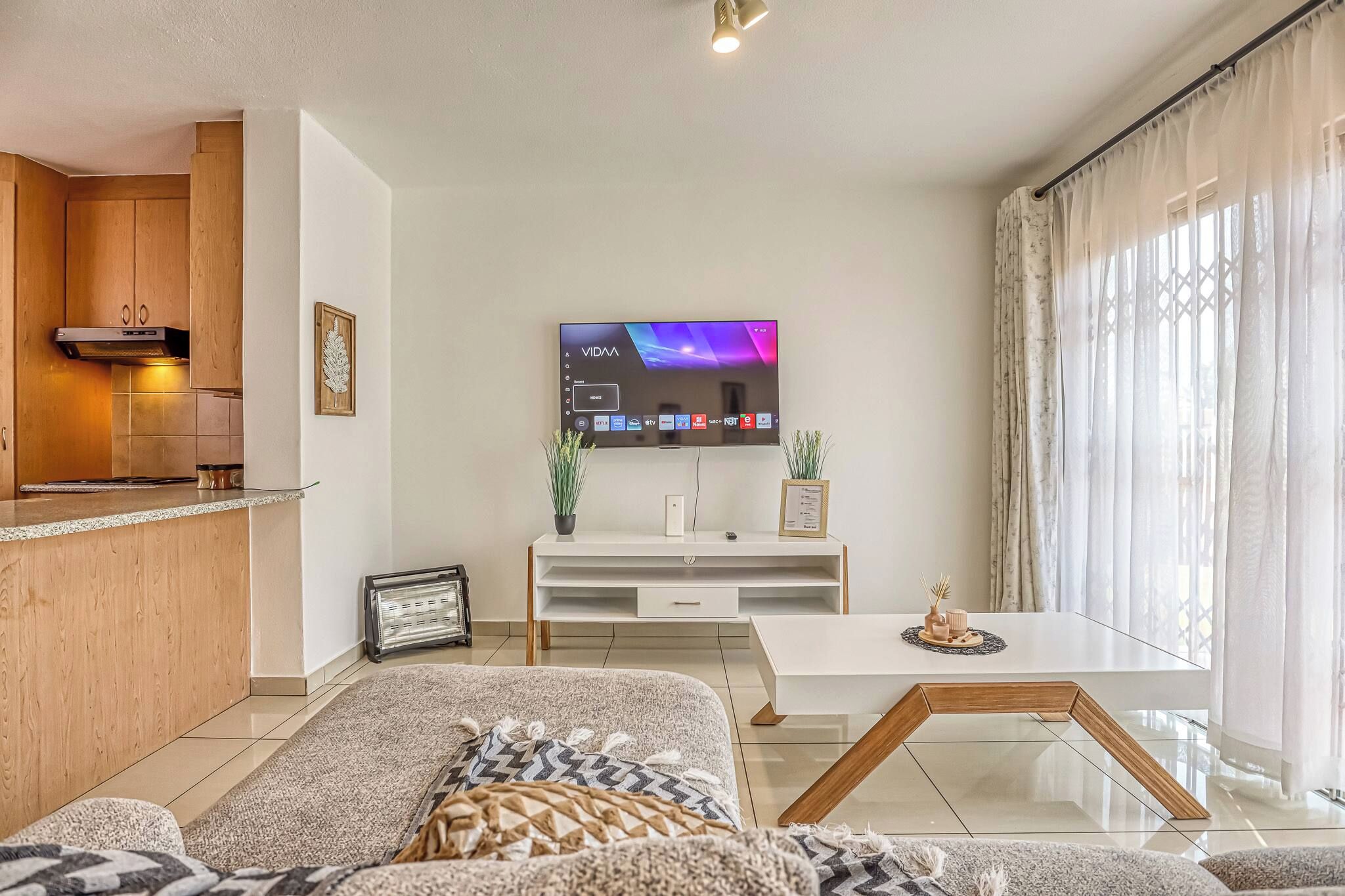 A modern living room with a flat-screen TV, a sofa, a coffee table, and a kitchen area with wooden cabinets and a built-in stove.
