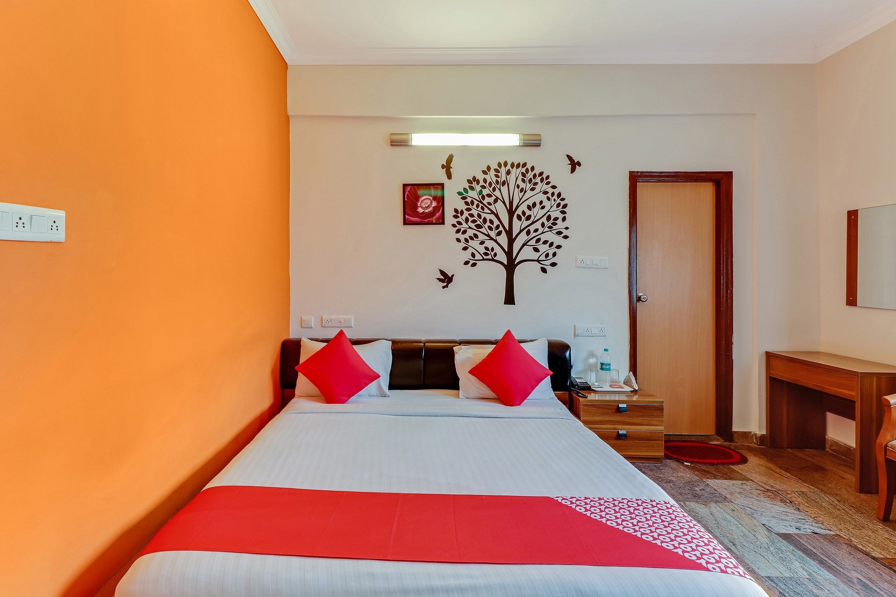 A hotel room with a bed, a desk, a chair, a door, and a wall with a tree decal.