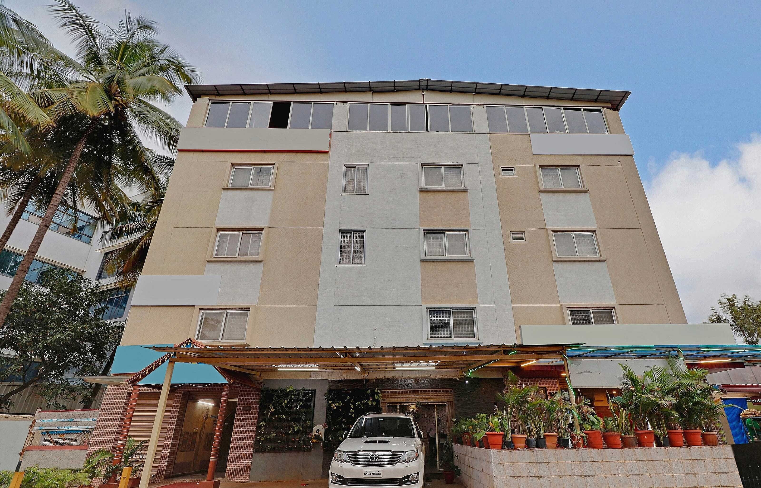 Un hotel de varios pisos con una entrada central, un coche estacionado en frente y una variedad de plantas en macetas alrededor del acceso.