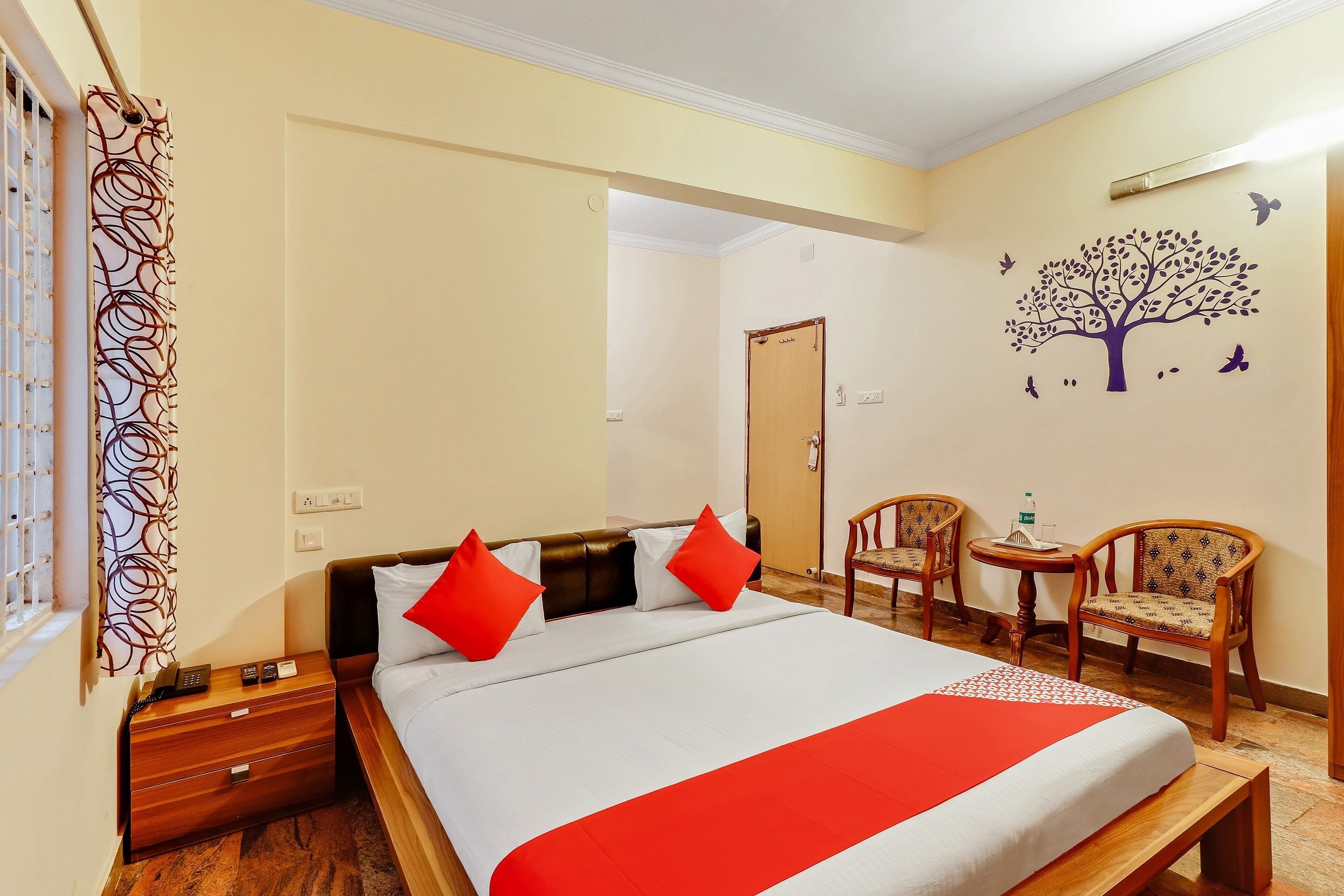 Habitación de hotel con cama, mesita de noche, lámpara y una decoración de árbol con pájaros.